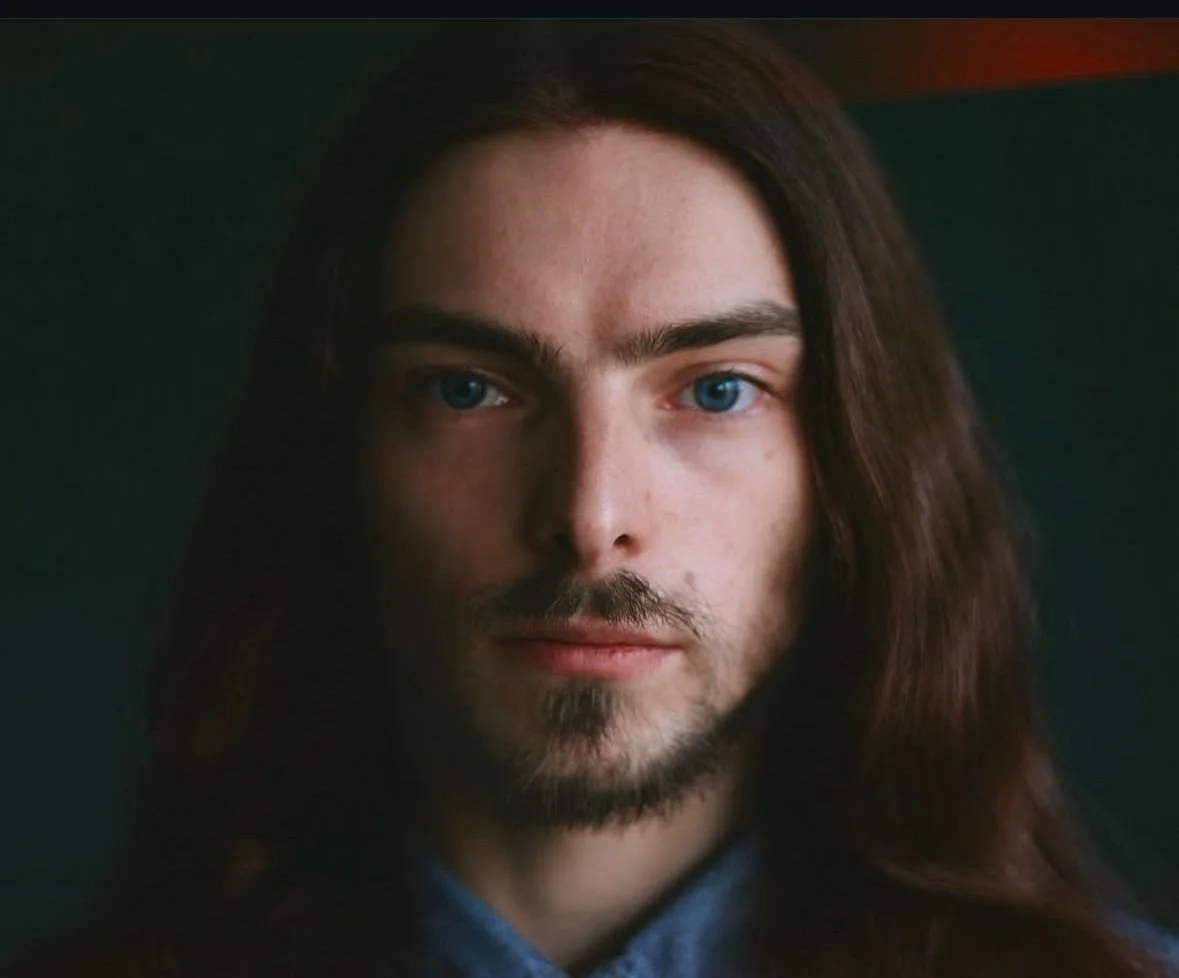 Close-up of a young man with long brown hair, blue eyes, light facial hair, and a neutral expression.