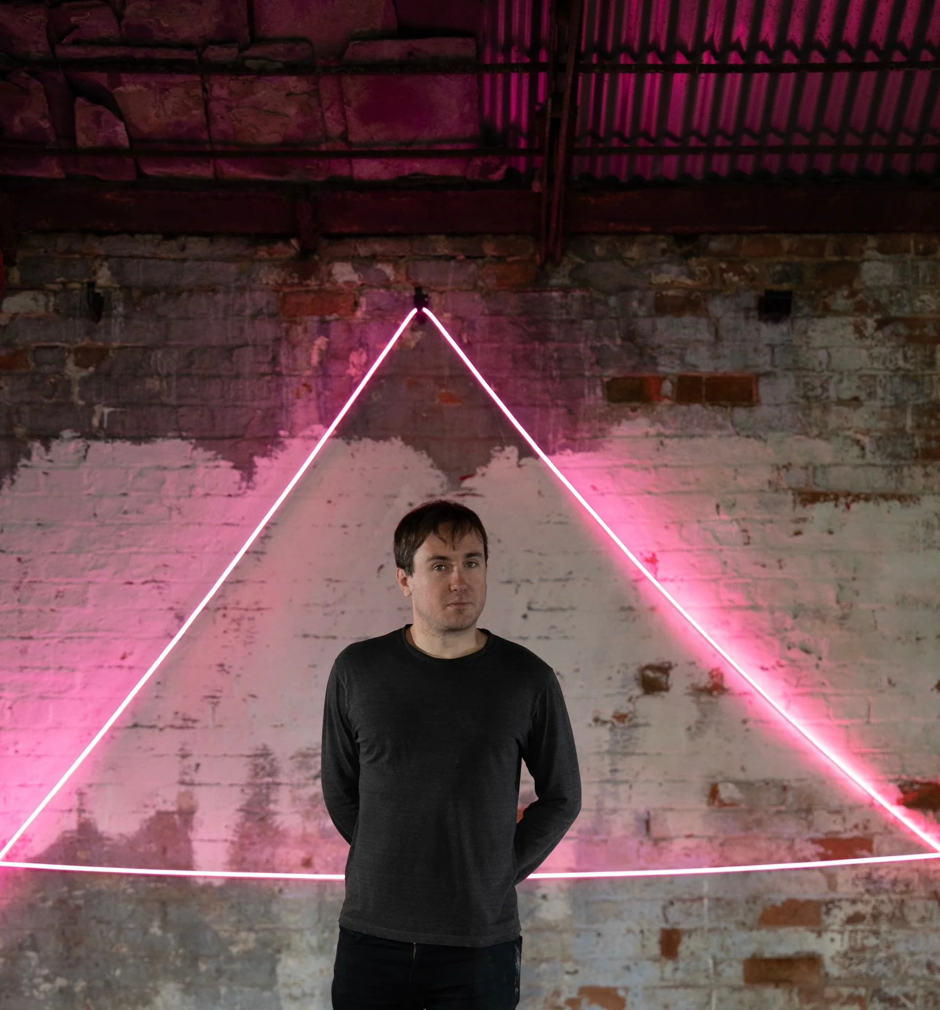 A man in a black shirt standing in front of a brick wall with a pink neon triangle light decoration behind him.