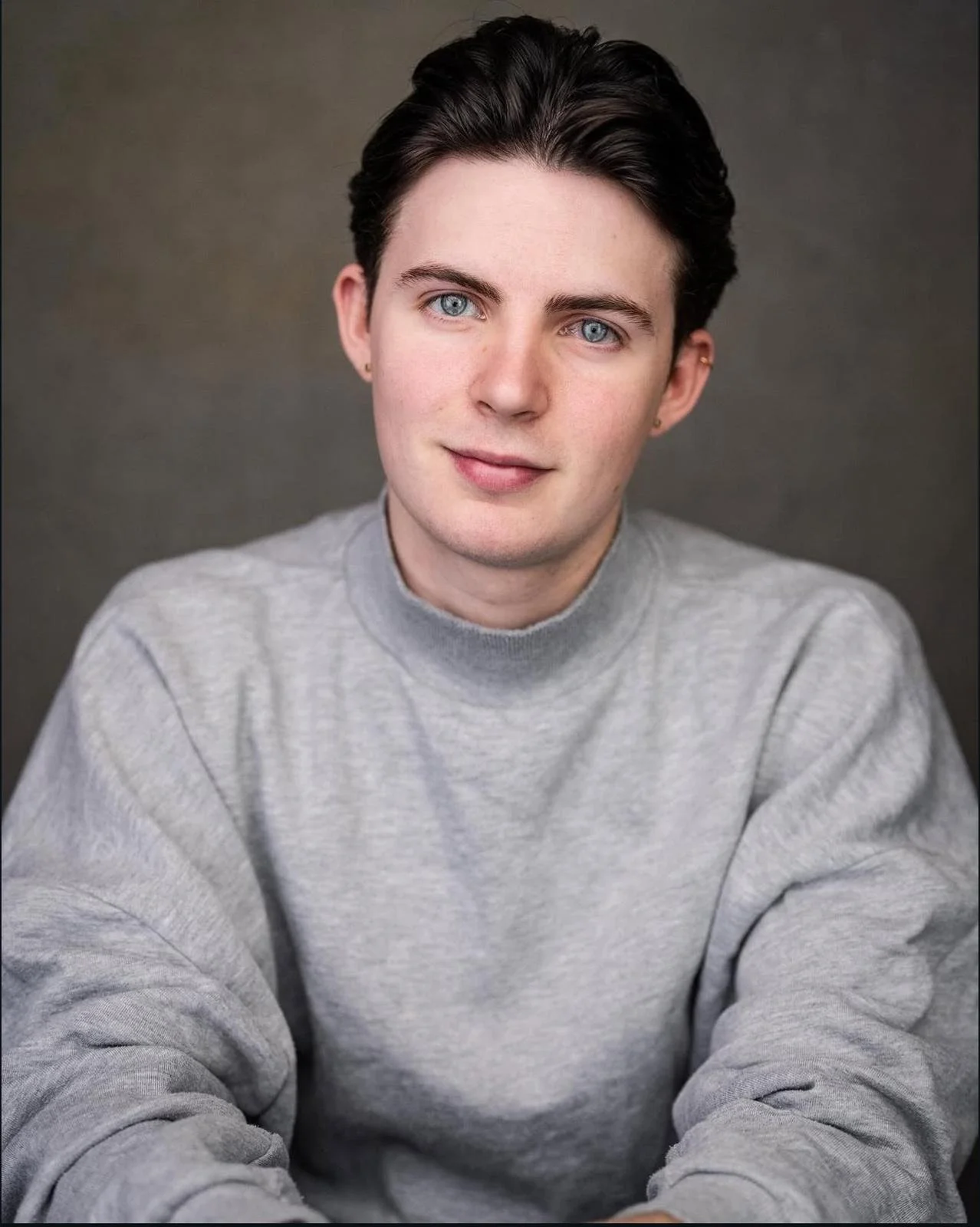 A young man with dark hair, blue eyes, wearing a gray sweatshirt, smiling at the camera against a dark gray background.