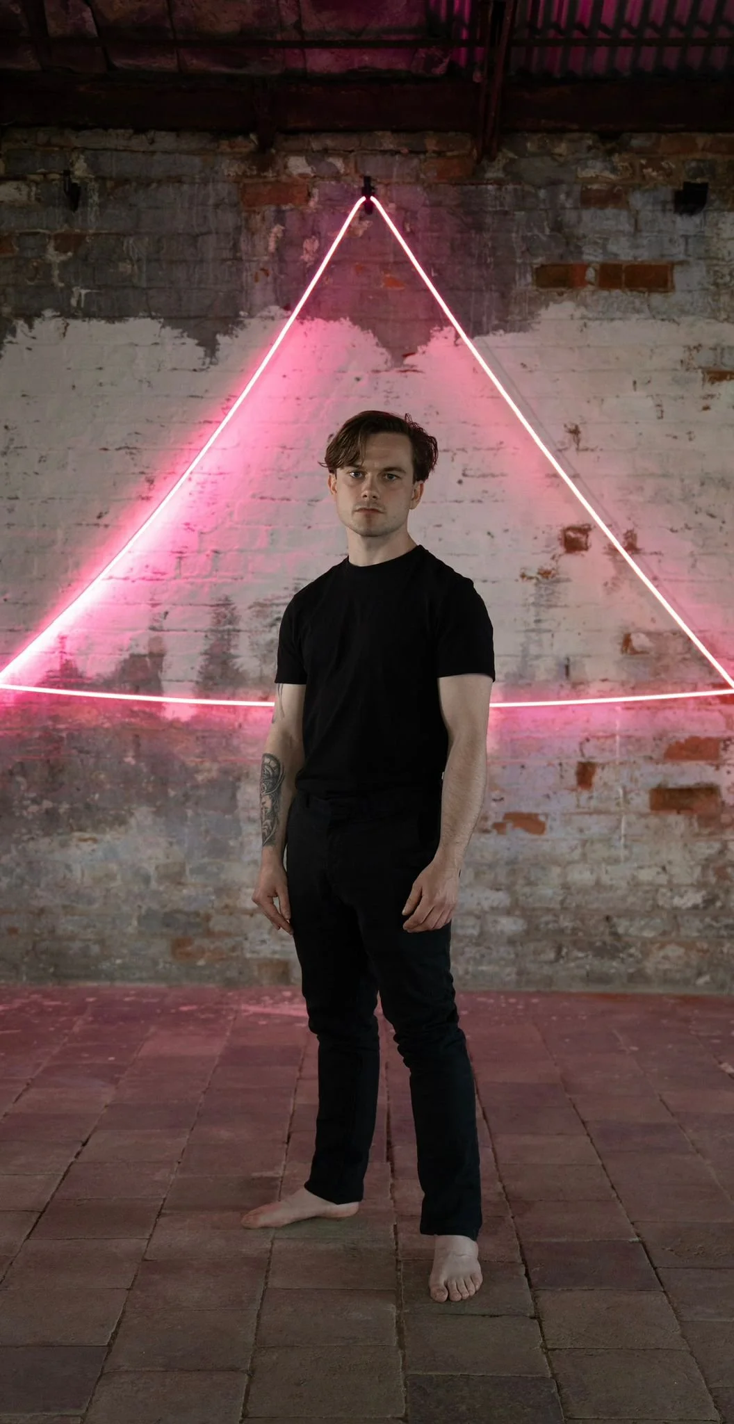 A young man with a tattoo on his right arm stands barefoot on a brick floor, wearing a black T-shirt and black pants, in front of a brick wall with pink neon light art arranged in a triangular shape.