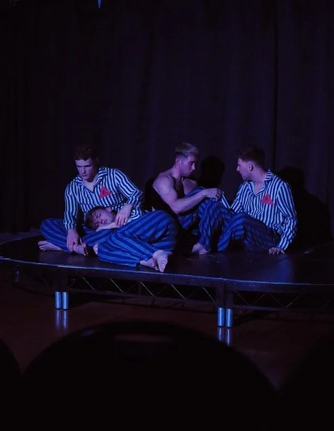 Three young men on a small stage, one lying down and two sitting, with dark curtains in the background. The two sitting men are wearing striped pajamas, and the man lying down appears to be asleep or resting.