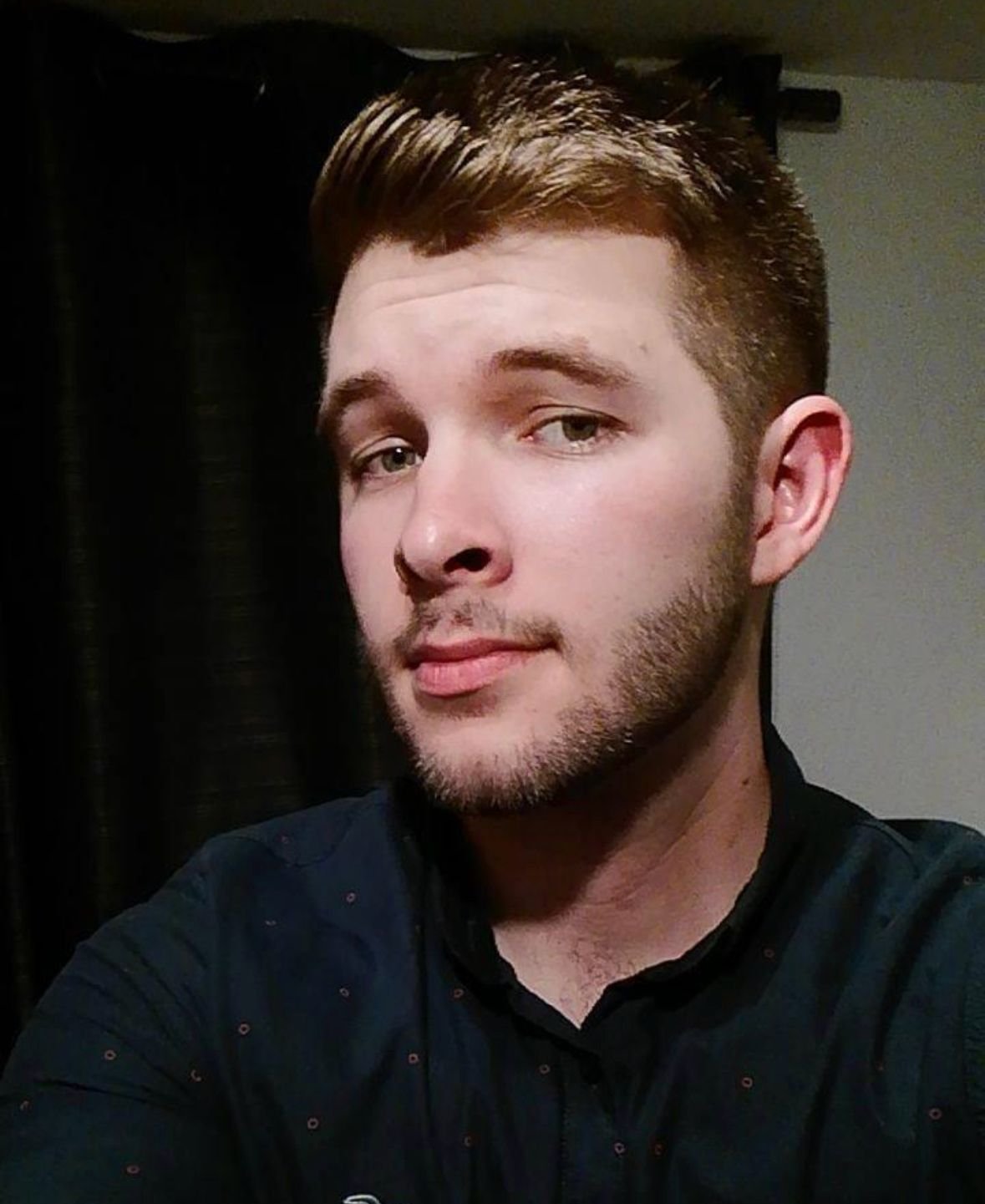 Close-up of a young man with short brown hair and a beard, wearing a dark shirt, looking at the camera with a subtle smile.