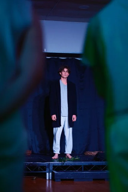 A young man standing on a small stage or platform, framed by blurred figures in the foreground, with a dark curtain behind him, in an indoor setting.