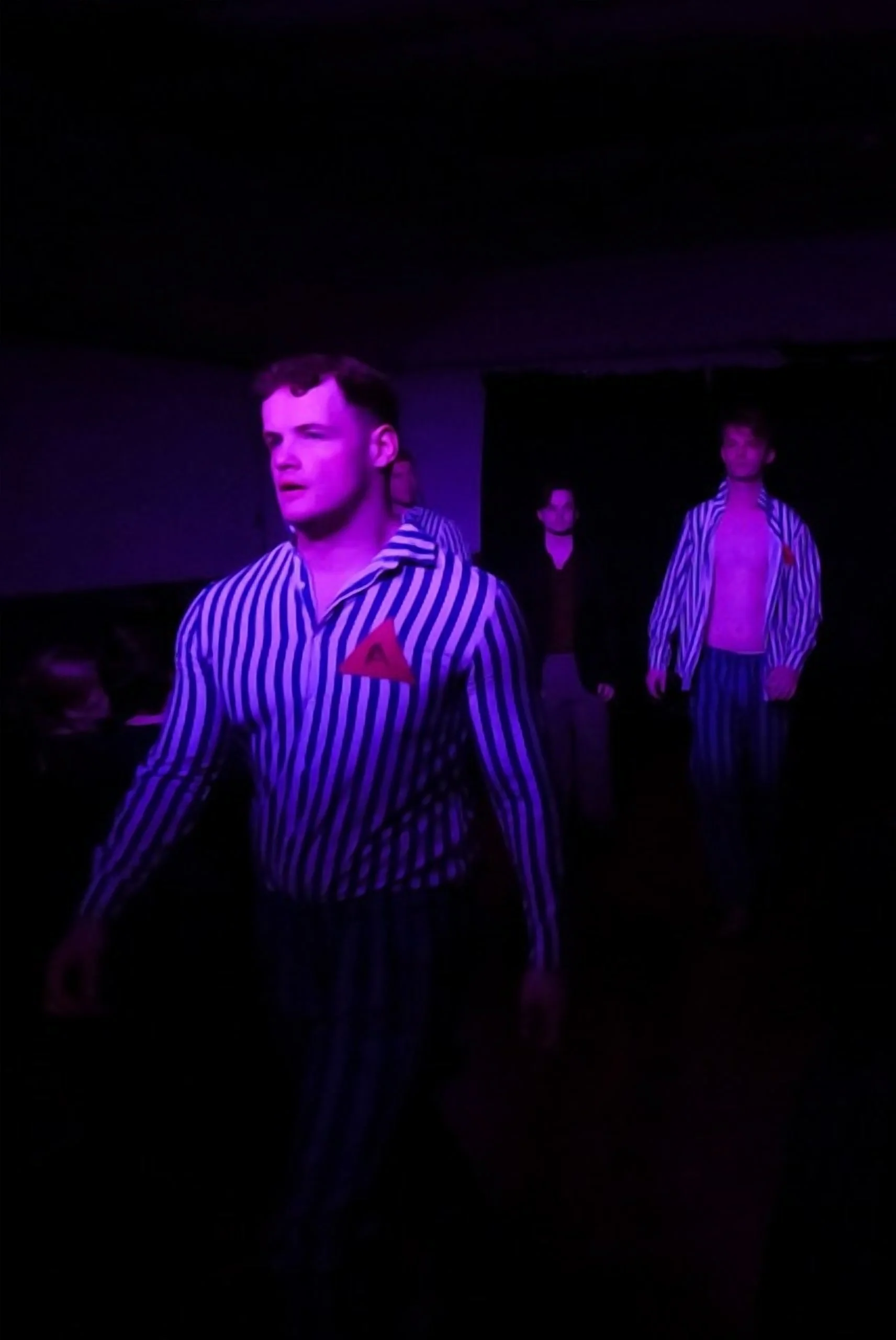 Actors in striped costumes on a dark stage, illuminated by purple lighting, participating in a theatrical performance.