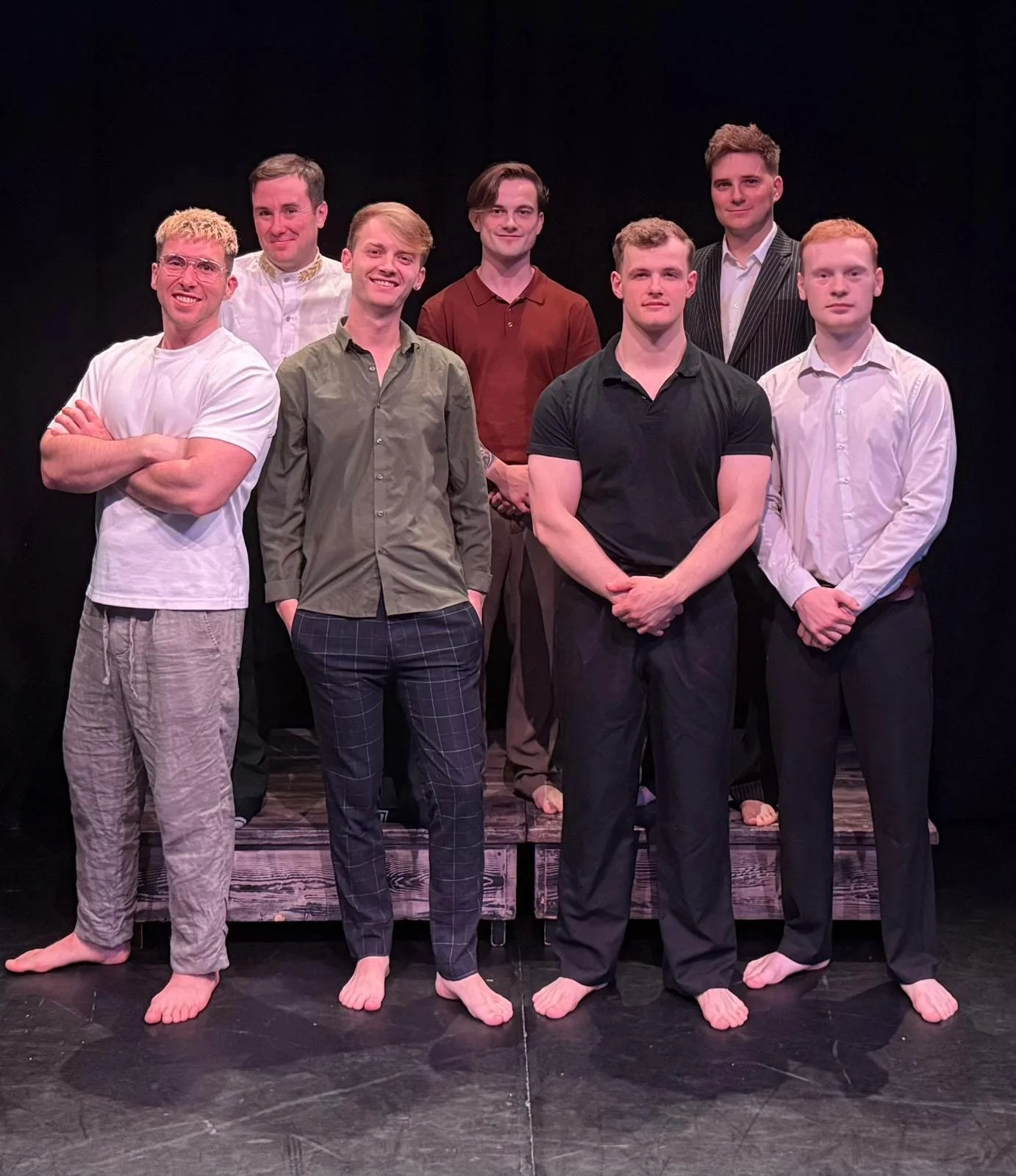 Group of nine men standing on a stage, some with arms crossed, dressed in casual and semi-formal clothing, barefoot, with a dark background.