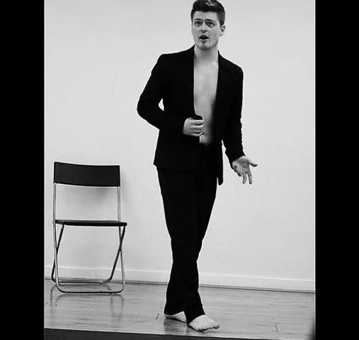 A black and white photo of a young man with short hair, partially dressed in a black blazer and pants, walking barefoot on a wooden floor, with a chair against the wall in the background.
