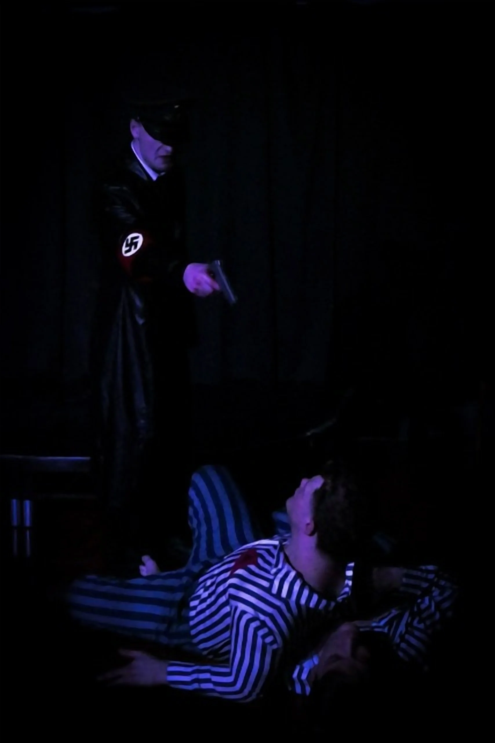 A person dressed in a Nazi uniform with a swastika armband holding a gun, standing in a dark room, looking downward at another person laying on the ground in striped pajamas, with blue and purple lighting.