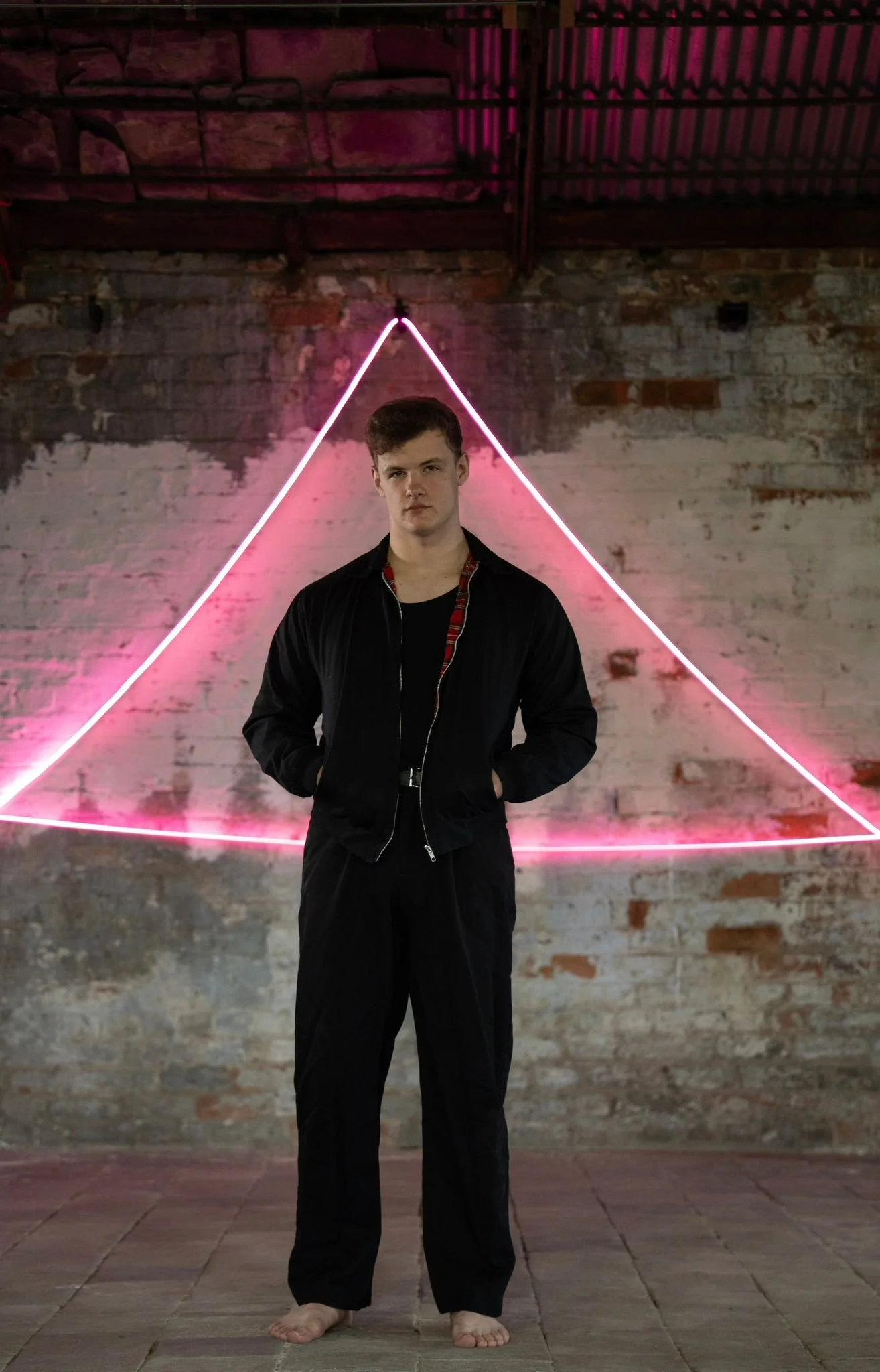 A young man standing barefoot in front of a brick wall with neon pink triangles, dressed in black pants and a black jacket, in an industrial-style space.