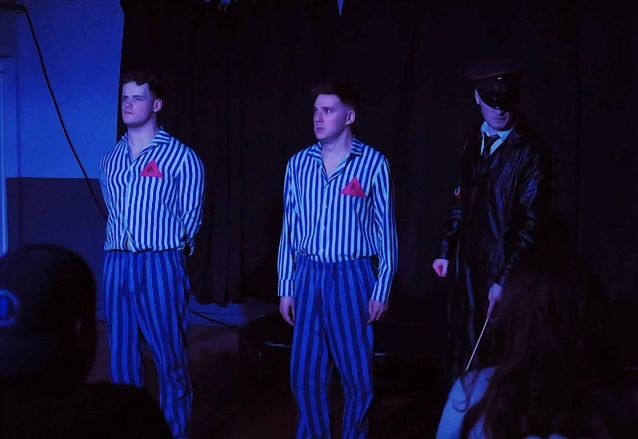 Two young men dressed in blue and white striped pajamas with red triangle insignias stand on a dark stage, with a third person dressed in black with a black leather jacket and a police-style cap. The scene appears to be part of a theatrical performan