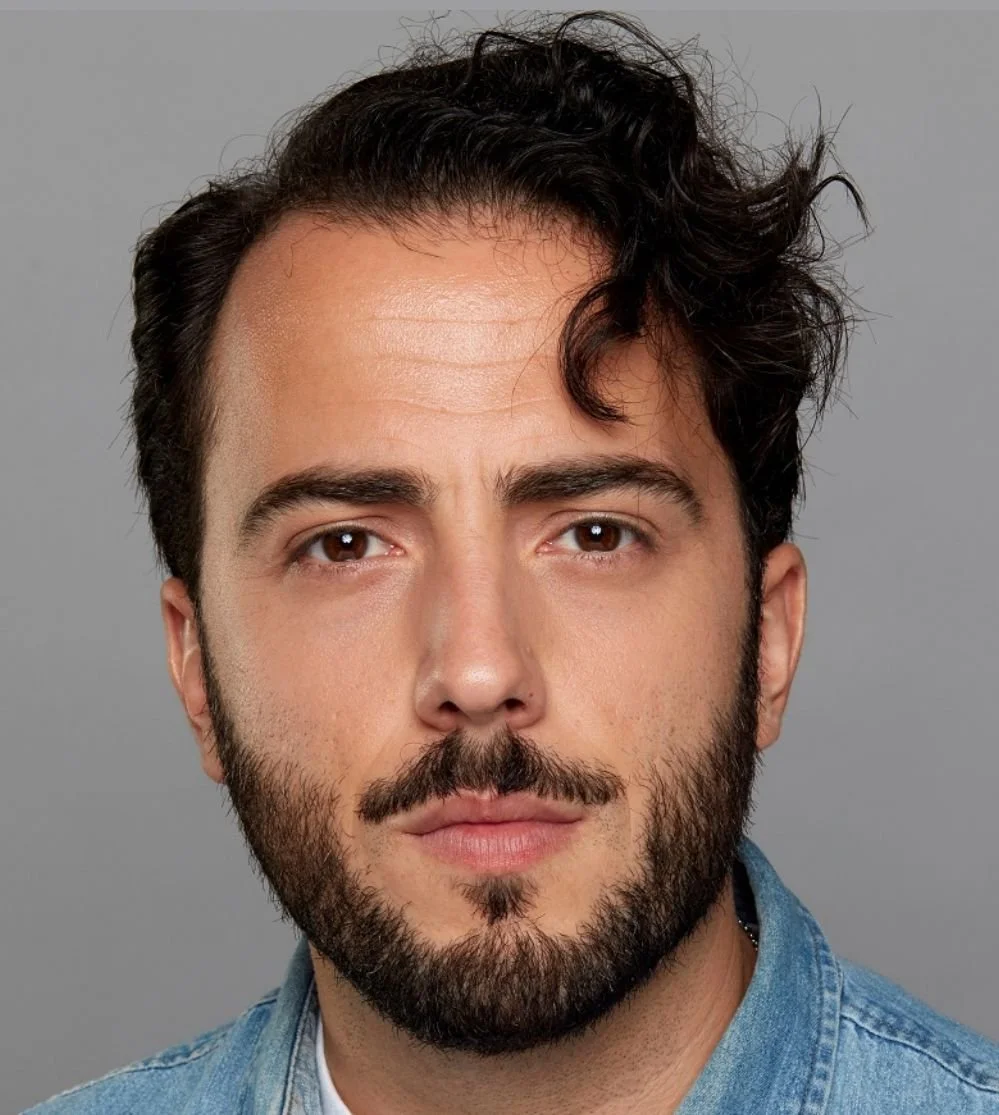 Close-up portrait of a young man with dark, wavy hair, facial hair, and wearing a denim shirt against a plain gray background.