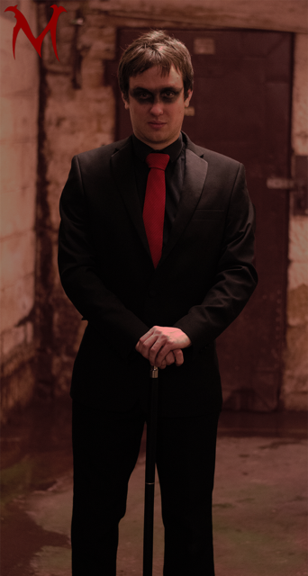 A young man in a black suit with a red tie stands with hands clasped in front in a dimly lit, rustic interior. He has dark hair and dark eye makeup, giving a dramatic appearance.