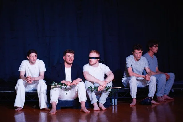 Five young men sitting on a stage, two with blindfolds, in what appears to be a theatrical performance or rehearsal.