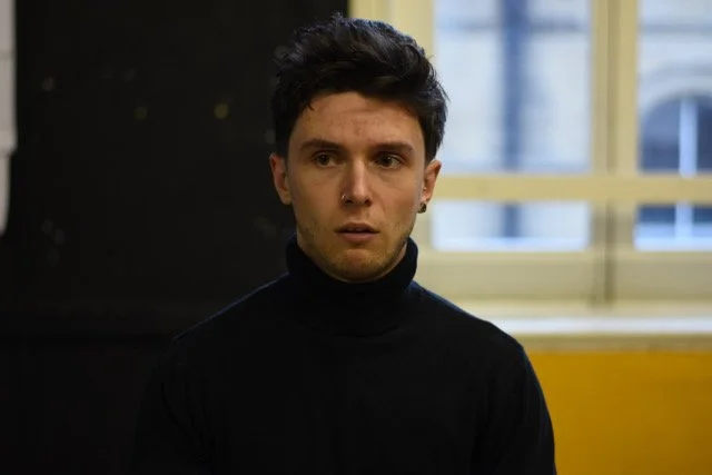 A young man with dark hair wearing a black turtleneck sweater, sitting indoors with a window in the background.