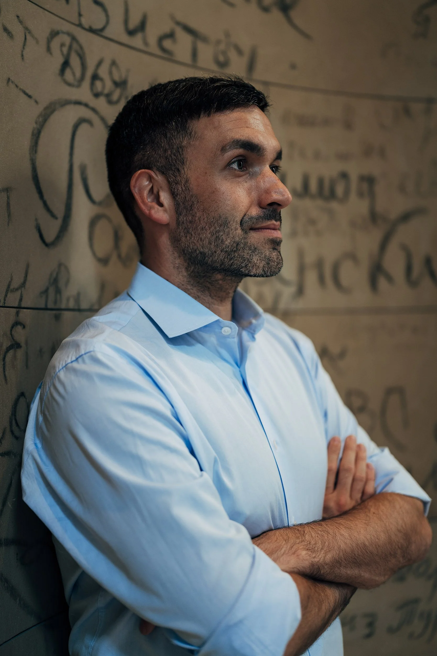 Ein Mann mit dunklen Haaren und Bart steht vor einer Wand mit schwarzen, handgeschriebenen Buchstaben, trägt ein hellblaues Hemd, hat die Arme verschränkt und schaut nach rechts.