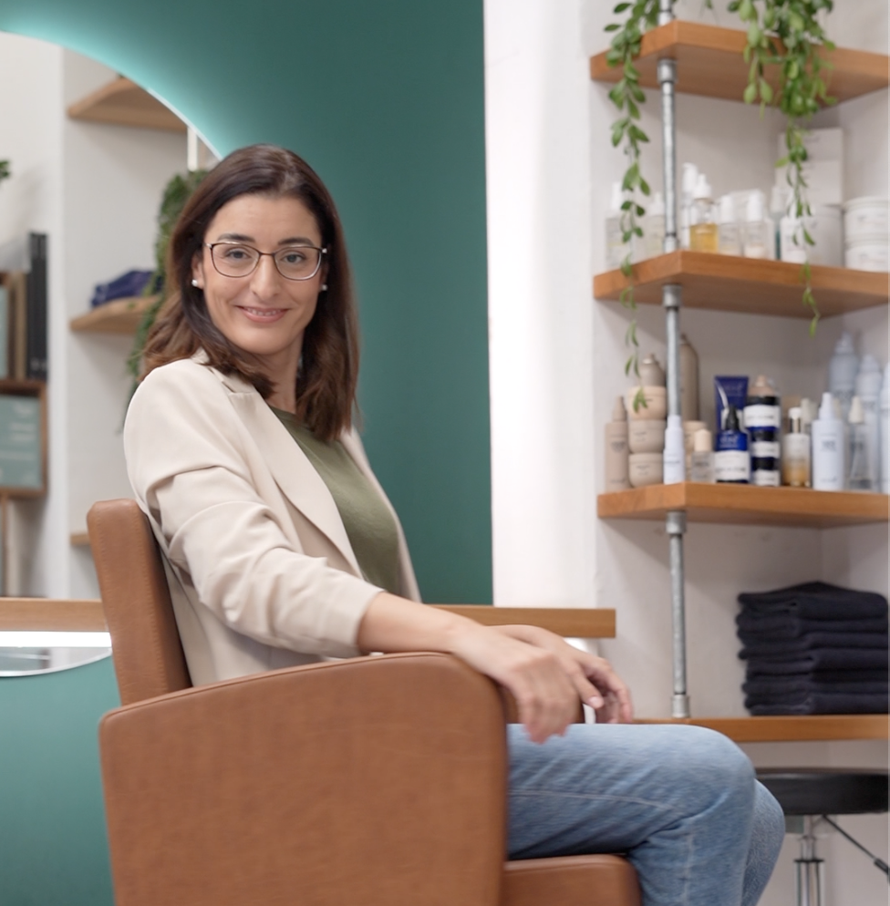 Eine freundlich wirkende junge Frau mit Brille sitzt in einem Kosmetikstudio oder Friseursalon und lächelt, während sie in einen bequemen braunen Stuhl sitzt. Im Hintergrund sind Regale mit Pflegeprodukten und Pflanzen zu sehen.