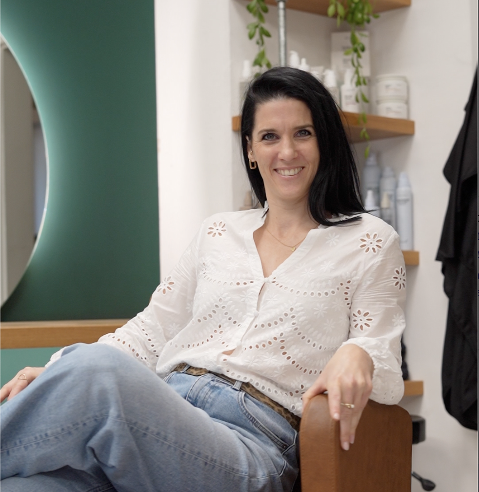 Eine lächelnde Frau mit schwarzen Haaren sitzt in einem Friseursalon.