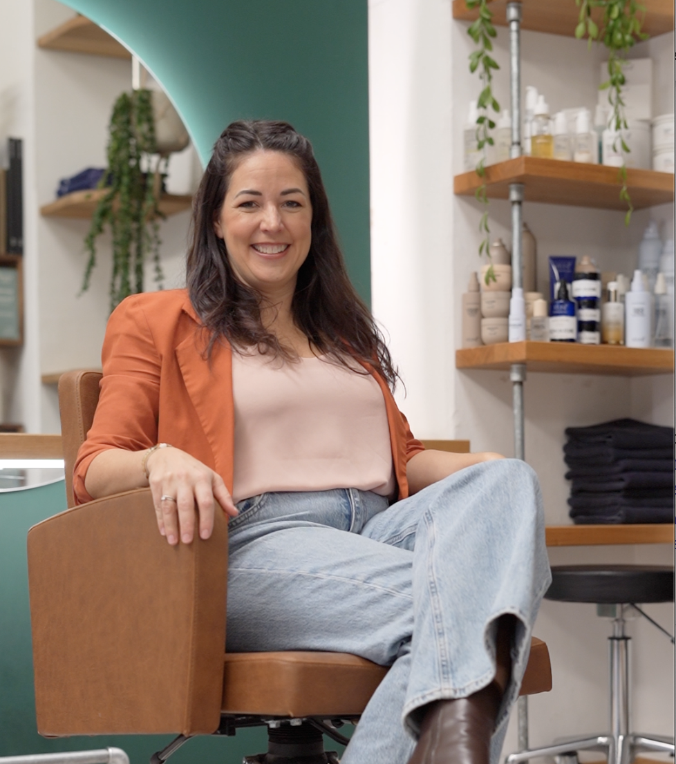 Eine lachende Frau sitzt bequem in einem Stuhl in einem Raum, der mit Regalen voller Kosmetik- und Pflegeprodukte sowie Pflanzen dekoriert ist.