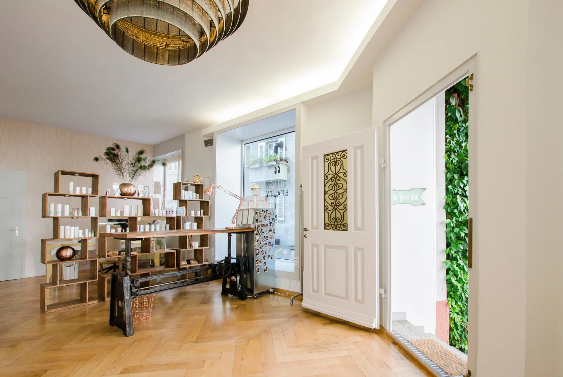 Ein heller Raum mit Holzboden, einer weißen Tür, einem Fenster und einem offenen Eingang mit grünen Pflanzen im Hintergrund. Es gibt eine Wandregalstruktur mit verschiedenen Dekorationsgegenständen, einem Tisch, einem Stehpult mit Fotos und einem Ste