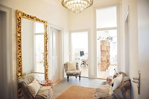 Ein helles Wohnzimmer mit einem großen, goldverzierten Spiegel, zwei antiken Sessel, einem kleinen Beistelltisch, einem Teppich und großen Fenstern, die den Raum mit natürlichem Licht erfüllen.