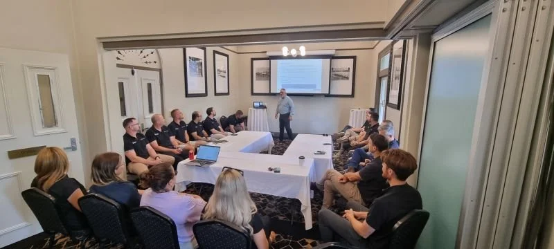 A man giving a presentation to a group of people seated in a conference room with a large monitor and framed pictures on the wall.