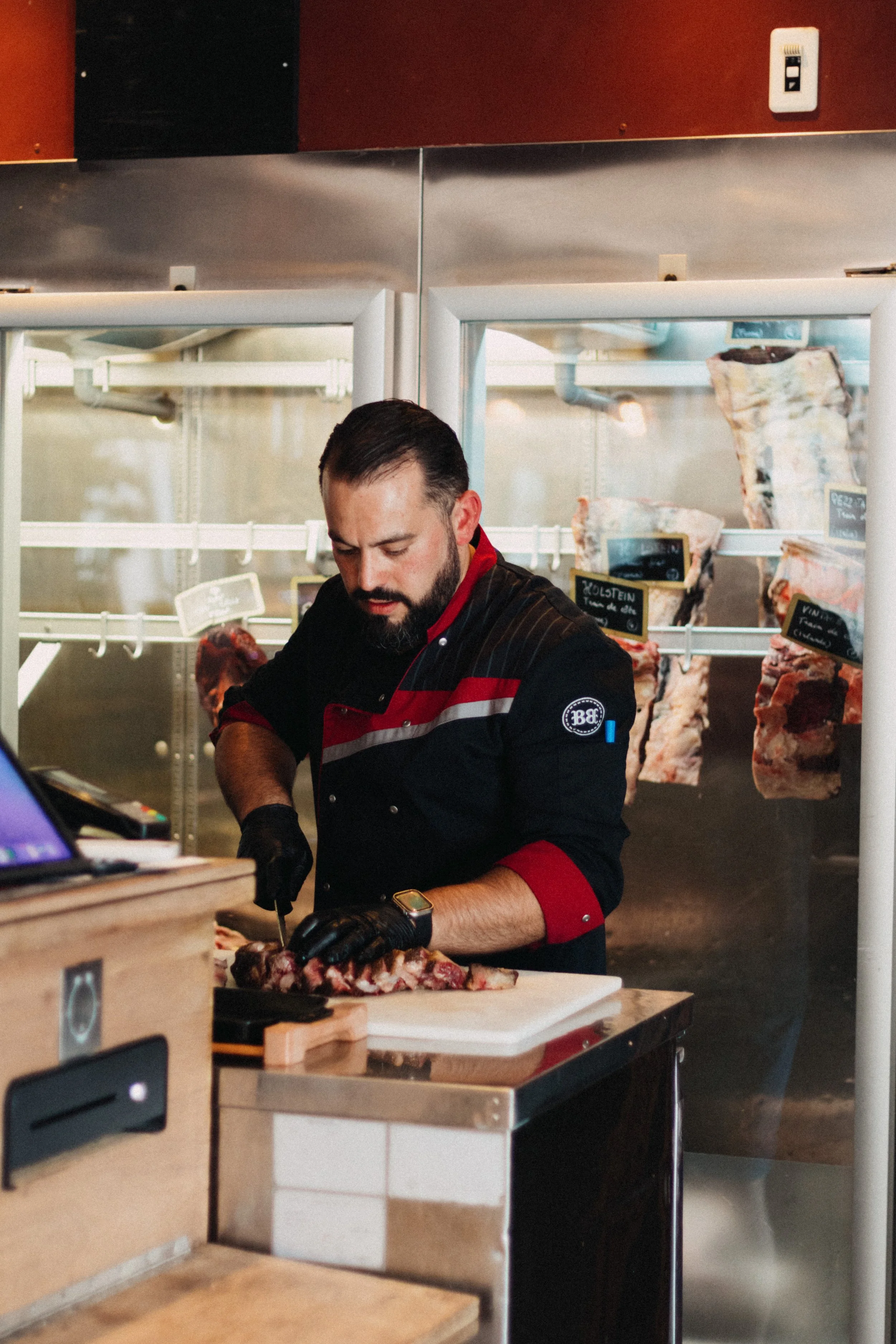 Un homme avec un uniforme de boucher, coupant de la viande sur une table en cuisine, avec de gros morceaux de viande suspendus en arrière-plan.