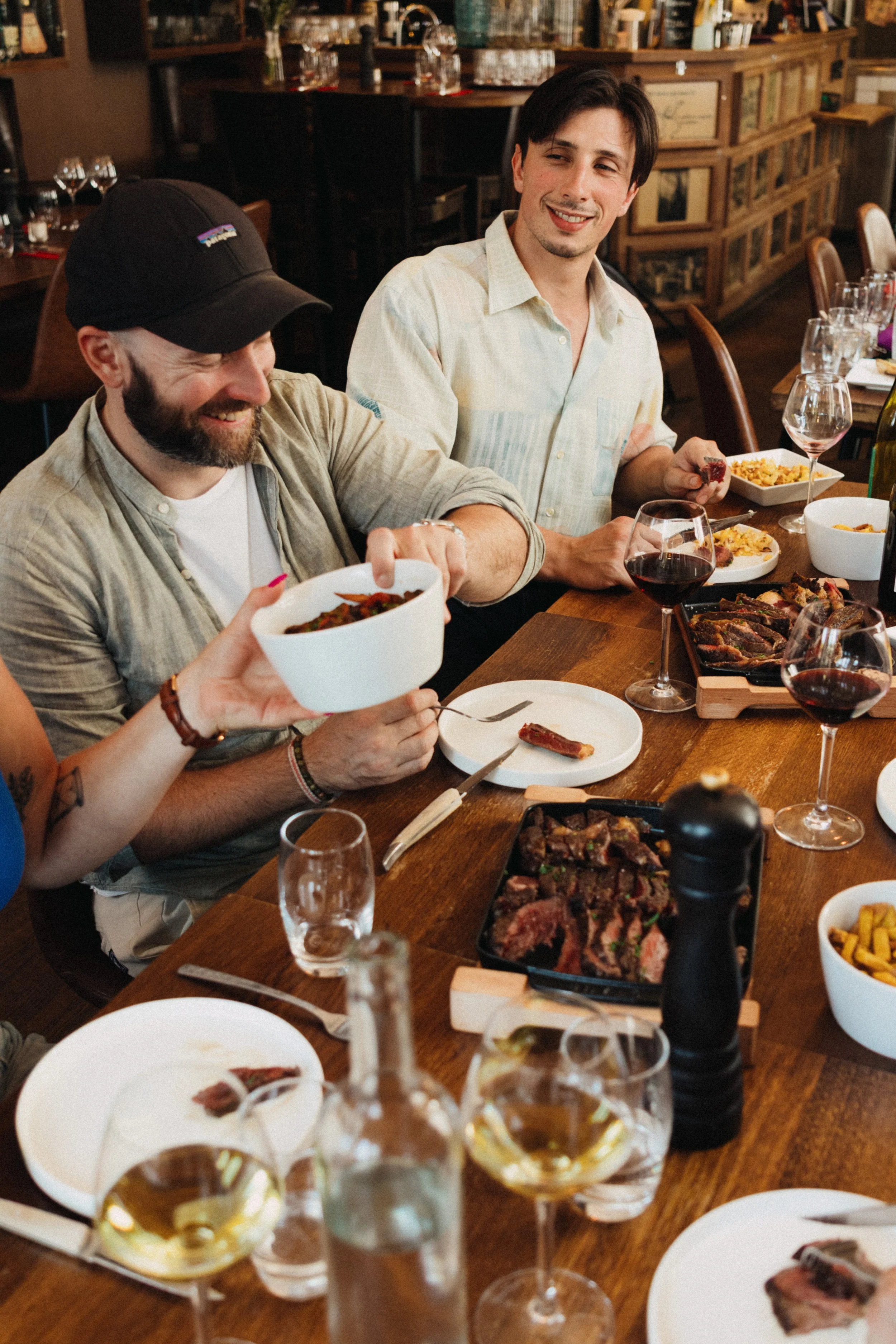 Une scène de repas convivial dans un restaurant avec deux hommes souriants partageant un moment autour d'une table garnie de plats variés, de verres de vin rouge, et de mets appétissants.