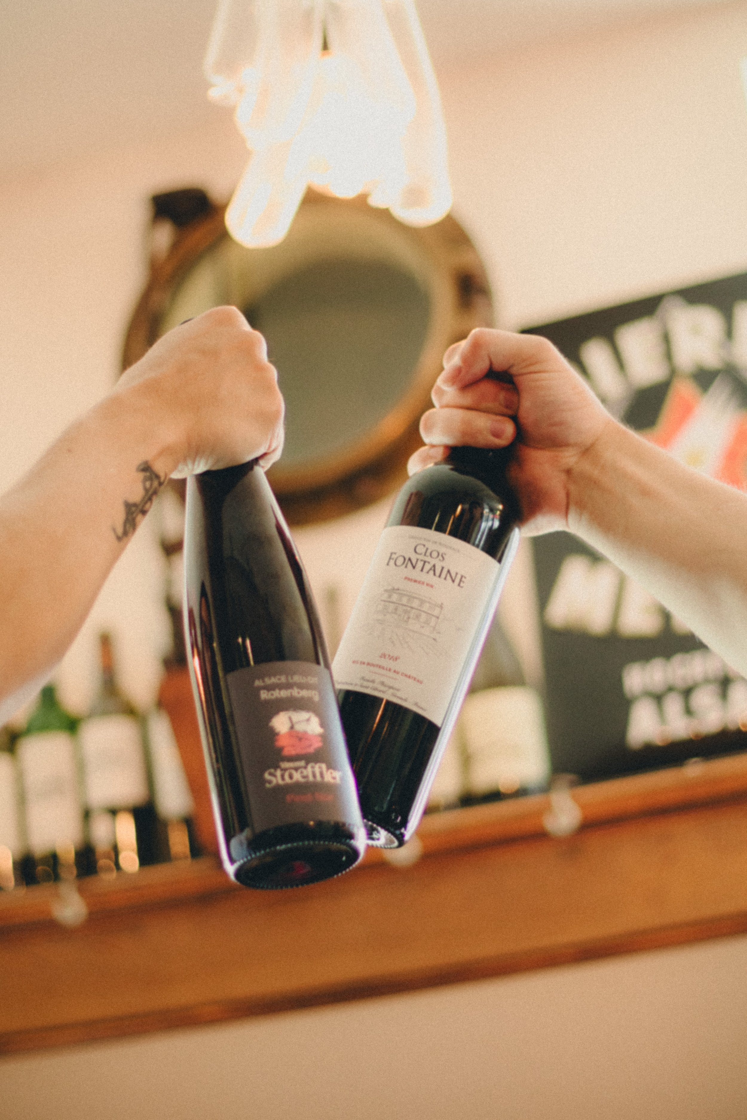 Deux mains tiennent des bouteilles de vin en plastique dans un environnement intérieur, avec un miroir rond en arrière-plan et un panneau noir avec du texte blanc, le tout dans une ambiance chaleureuse.