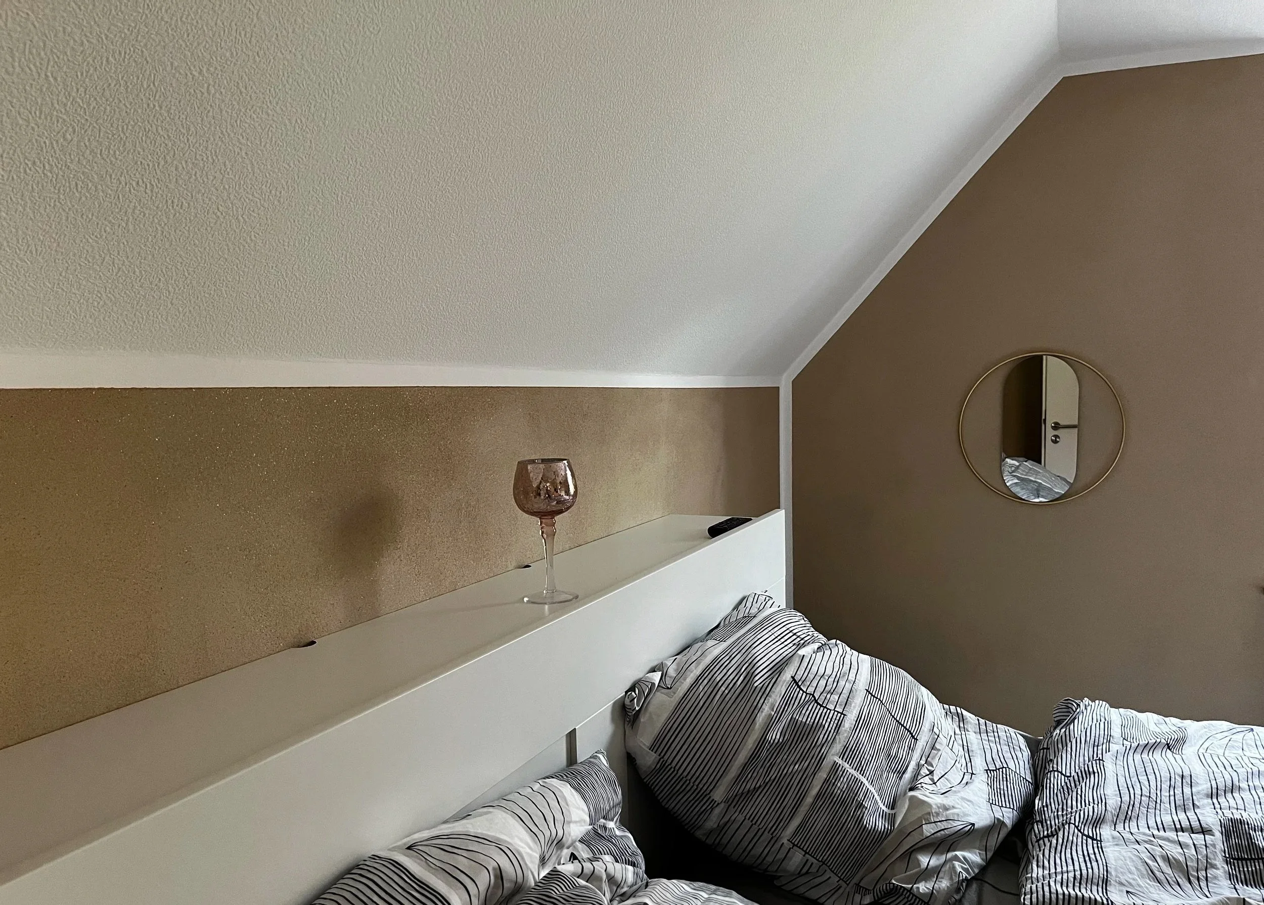 Ein Schlafzimmer mit einem Doppelbett, Gebrauchsspuren auf den Bettwäsche, ein runder Wandspiegel, ein Nachttisch mit einem Glas und einer Fernbedienung, weiße und braune Wände, eine geschrägte Decke.