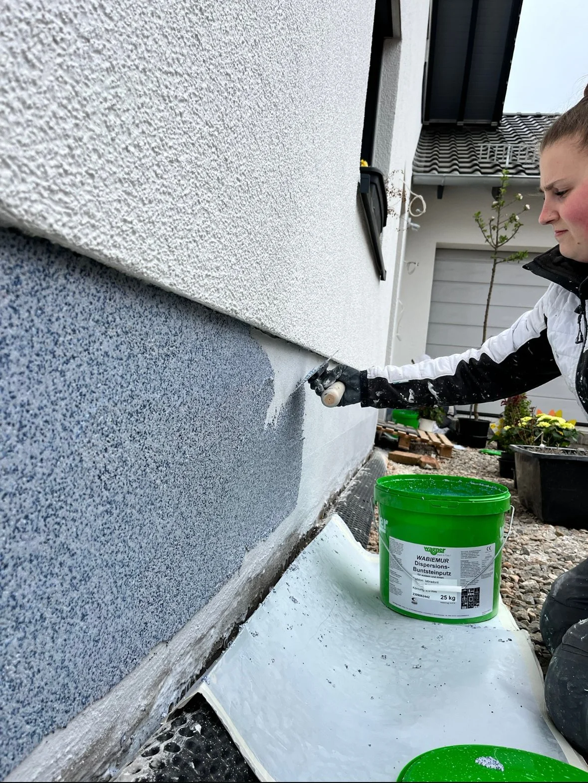 Person trägt weißen und schwarzen Mantel und arbeitet draußen an der Hauswand, beim Streichen oder Verputzen, mit einem grünen Eimer auf dem Boden.