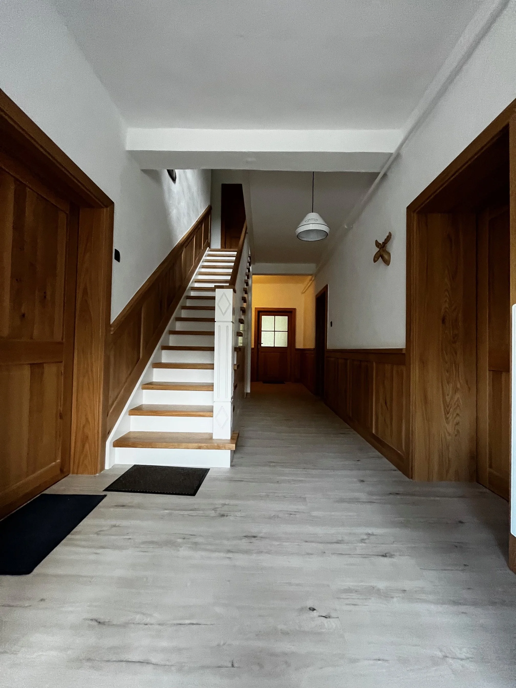 Innenansicht eines Flurs mit Treppe, Holzverkleidungen, weißem Boden und weißen Wänden, mit einer Deckenlampe und einer Wanddekoration.