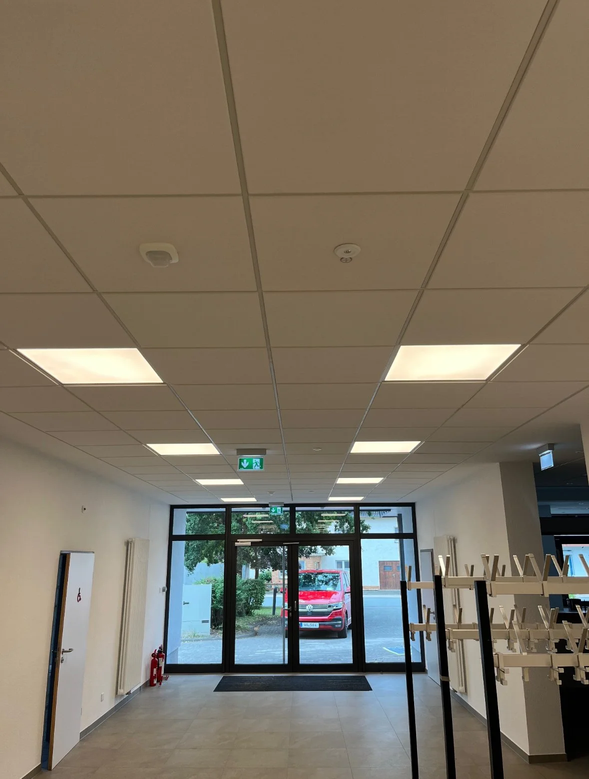 Innenaufnahme eines Eingangsbereichs mit Glastür, Wasserkocher, Brandschutz, und modernen Deckenleuchten.