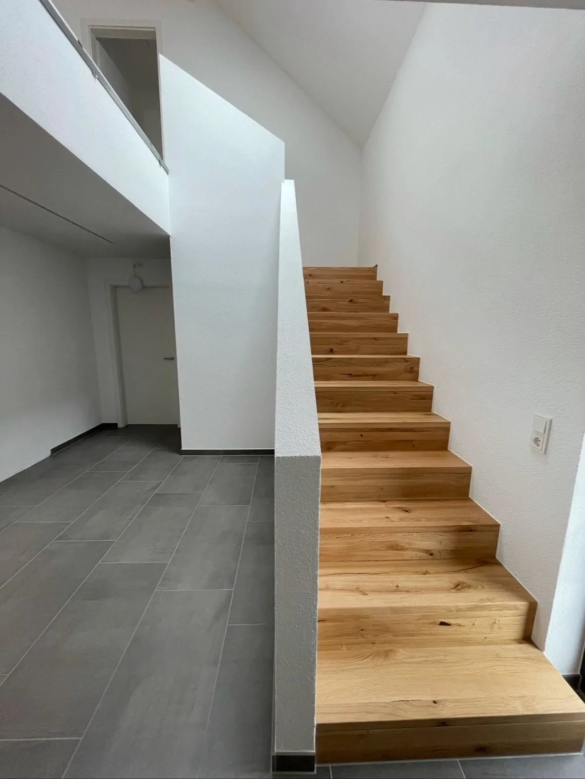 Innenansicht eines Hauses mit Treppe aus Holz, weiße Wände, graue Fliesenboden, Tür im Hintergrund.