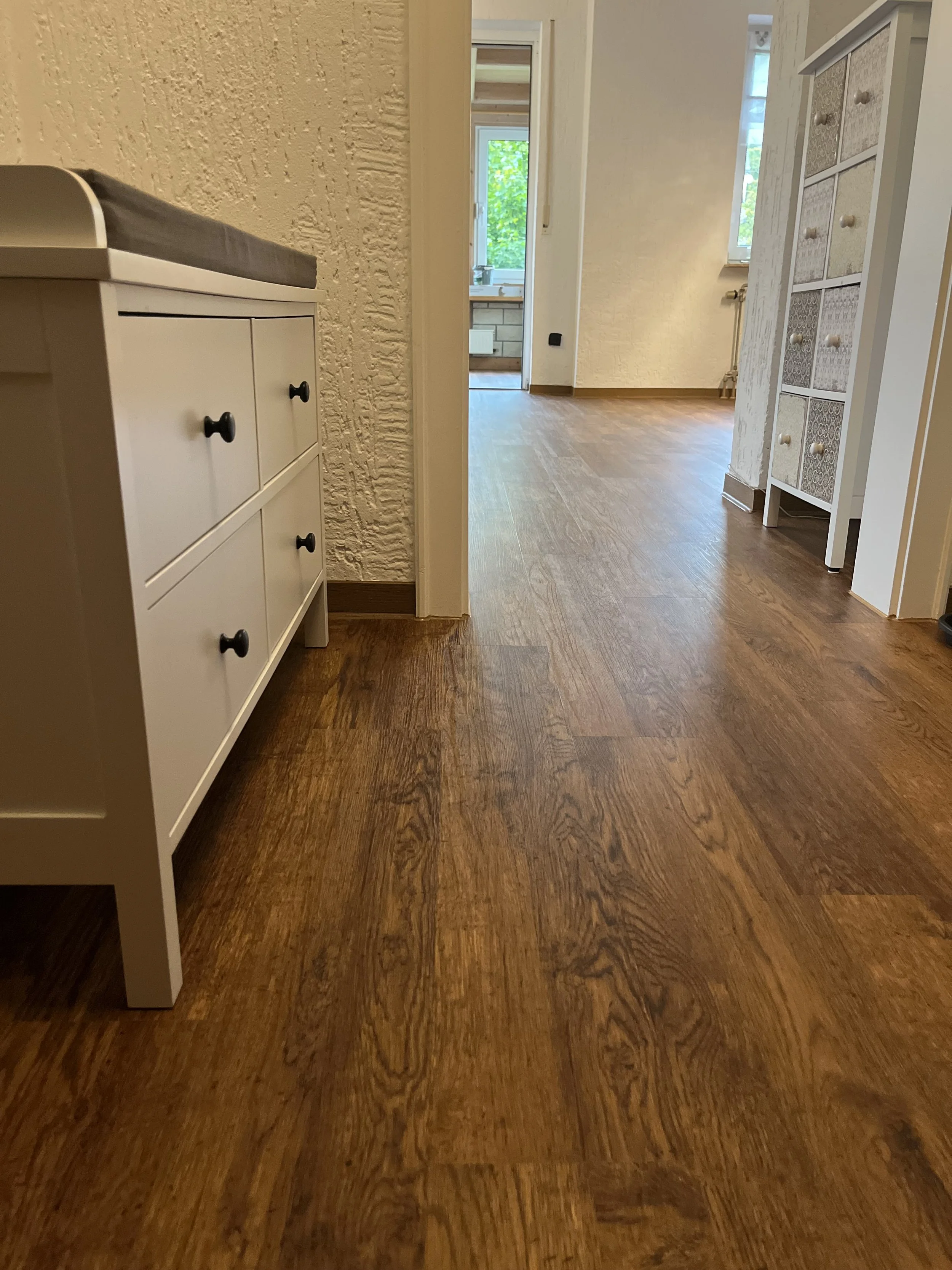 Bodenbelag aus Holzparkett in einem Flur mit weißen Möbeln und einem offenen Raum mit Fenstern im Hintergrund.