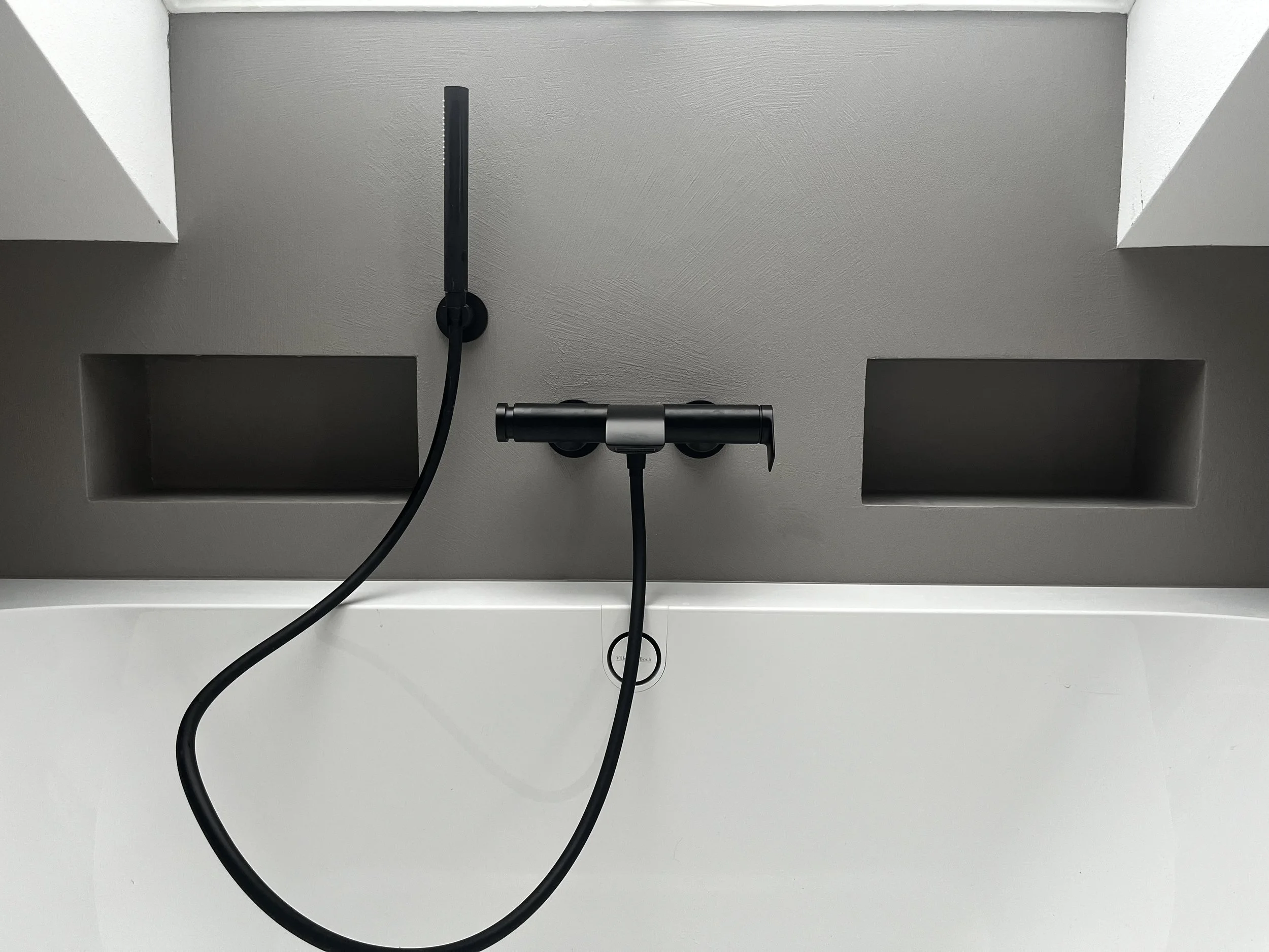 Wand mit zwei schwarzen Steckdosen, einem Wandmontierten schwarzen Handbrause und einem schwarzen Wasserhahn, über einer weißen Badewanne.