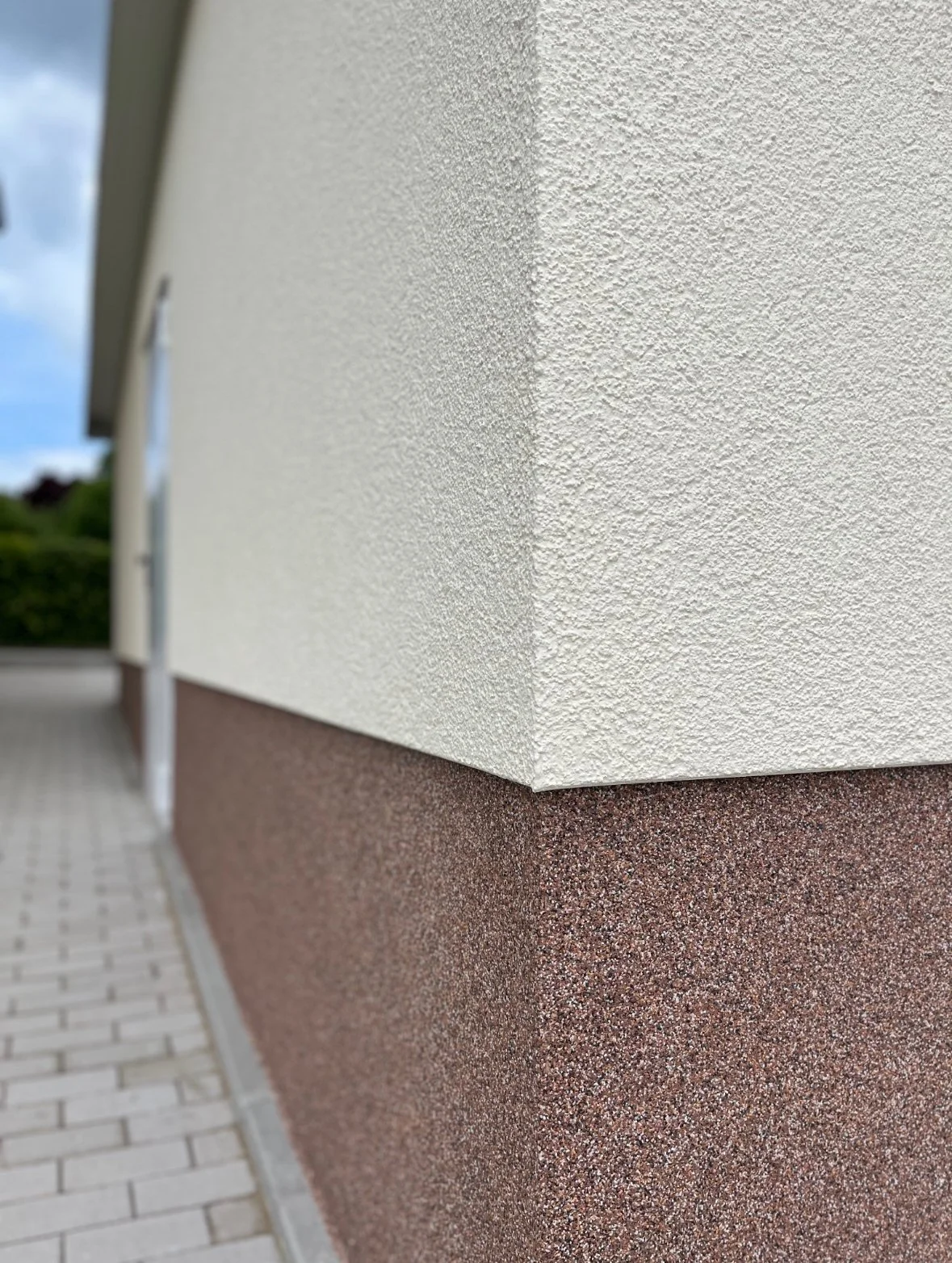 Ansicht eines Gebäudes mit weißer, strukturierter Außenwand und brauner Grundmauer, neben einem Gehweg aus Ziegelsteinen.