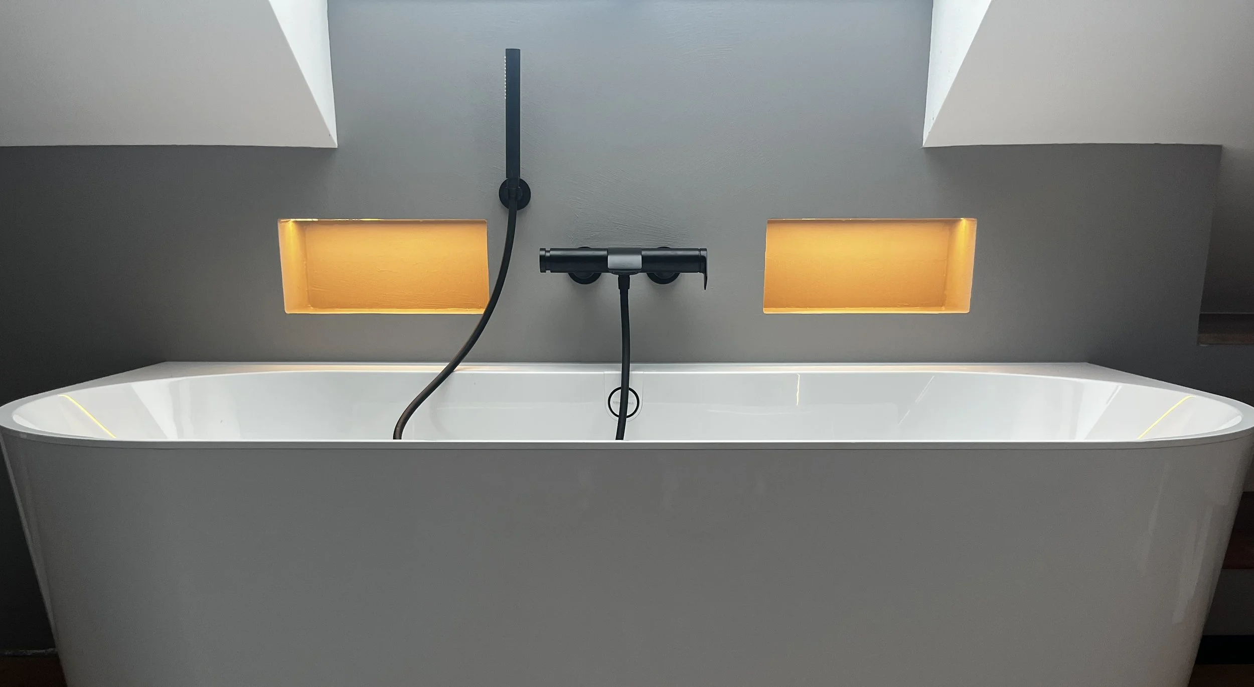 Moderne freistehende Badewanne mit hellrosa Innenraum, integrierter Badarmatur und beleuchteten Nischen in der Wand, mit Tageslicht durch Oberlichter