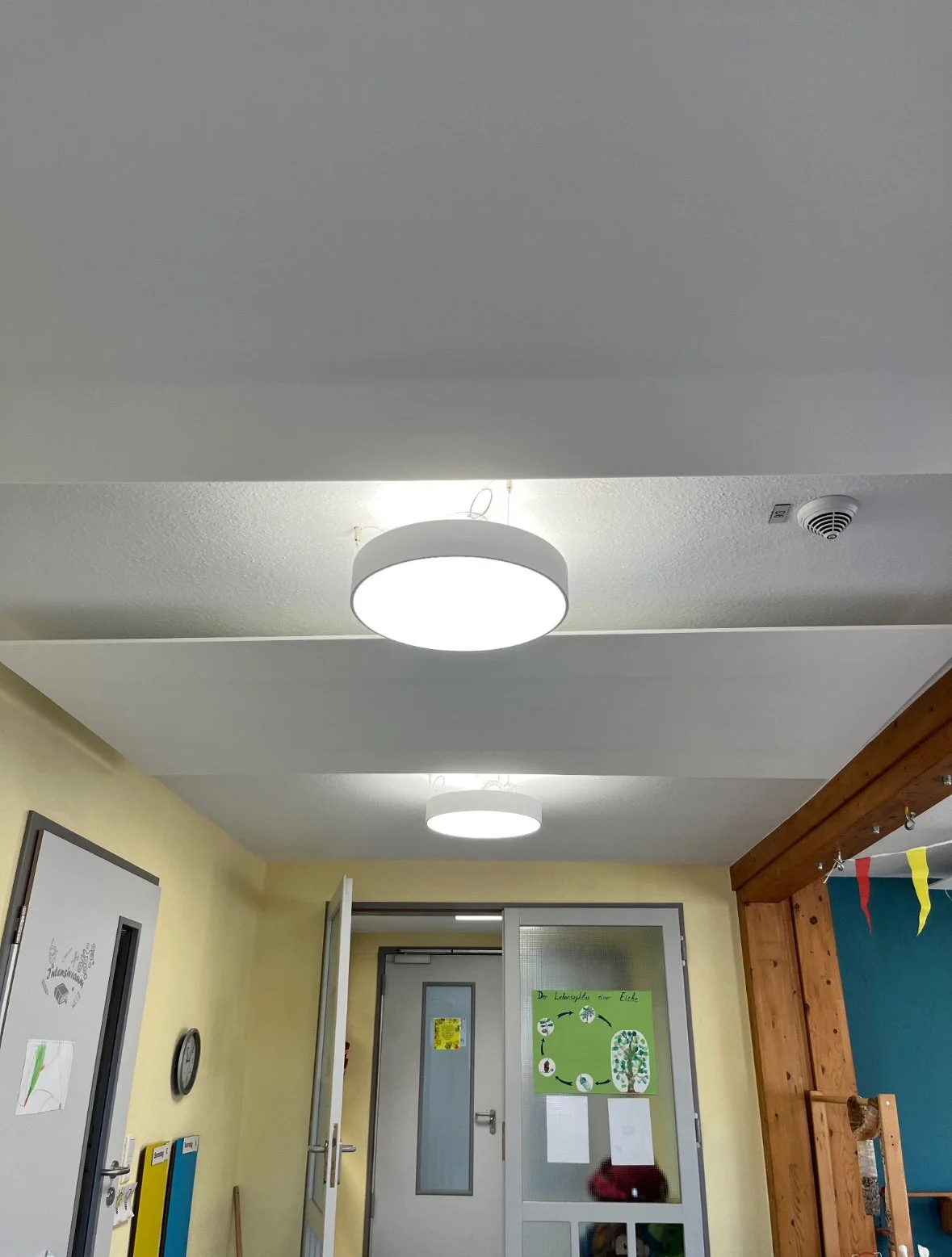 Bild von einem Flur in einem Gebäude mit weißen Deckenleuchten, einer gelben Wand, einer Tür, einem Wanduhr, und einem grünen Poster.