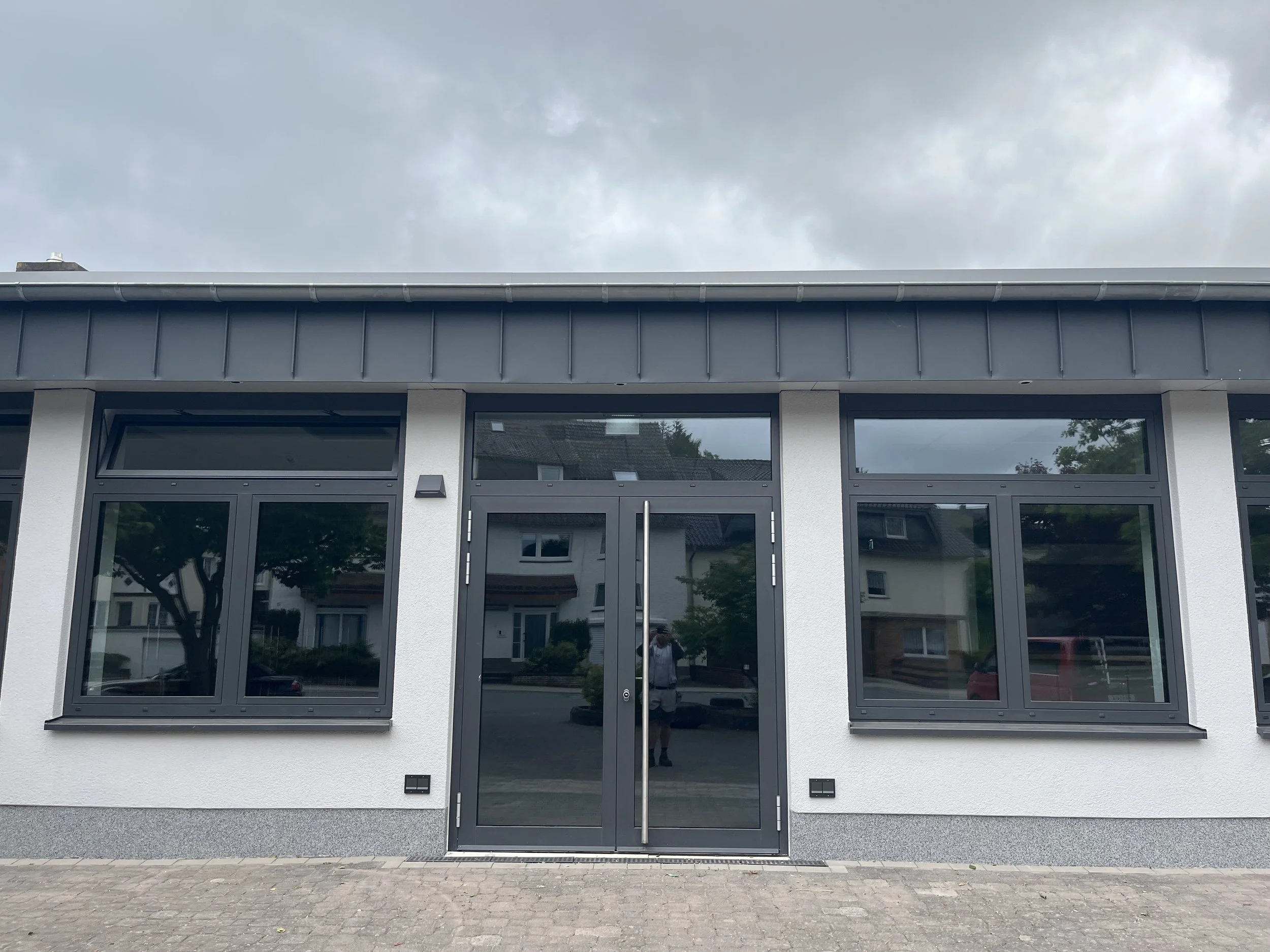 Außenansicht eines modernen Gebäudes mit großen Fenstern und einer schwarzen Tür, bei bewölktem Himmel.