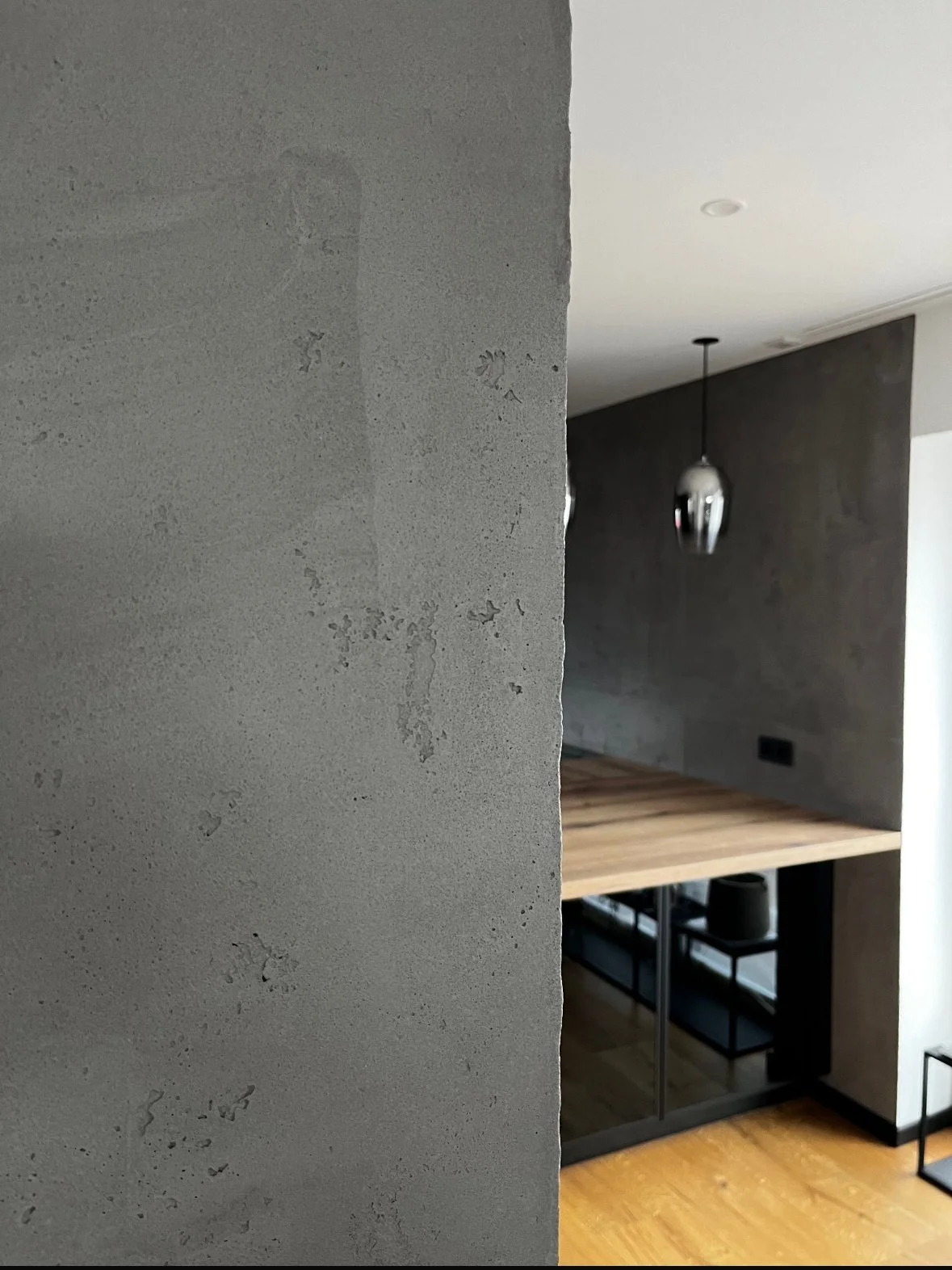 Innenaufnahme eines Raumes mit modernem Design, eine Wand aus rohem Beton, ein Küchenbereich mit Holzarbeitsplatte, schwarzem Unterschrank und einer pendelnden Lichterkugellampe.