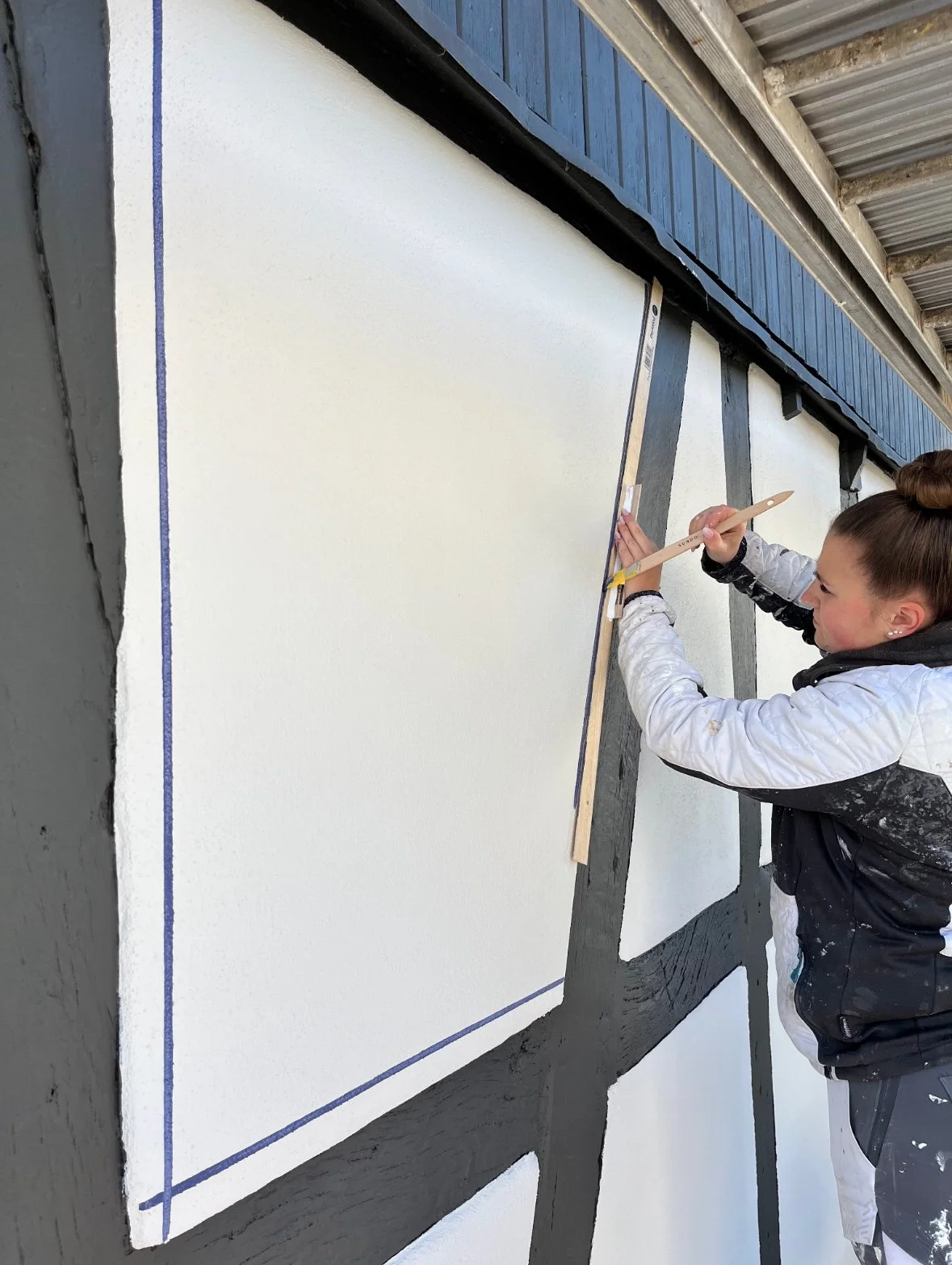 Junge Frau bemalt die Wand eines Fachwerkhauses mit einem Pinsel, während sie die Linien eines Designs nachzeichnet.