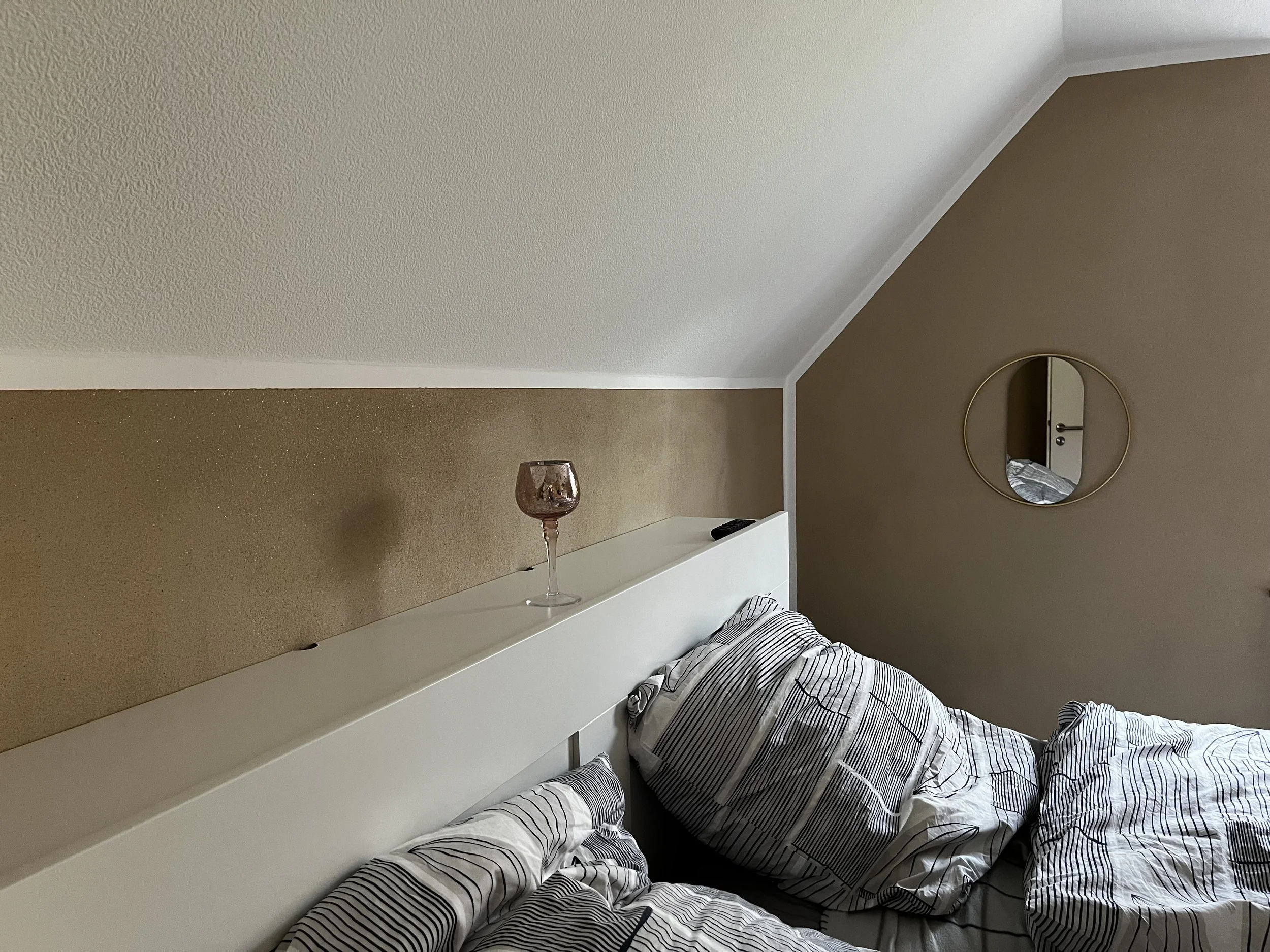 Ein Schlafzimmer mit unordentlichem Bett, einer Kerze auf einem weißen Nachttisch und einem runden Spiegel an der Wand.