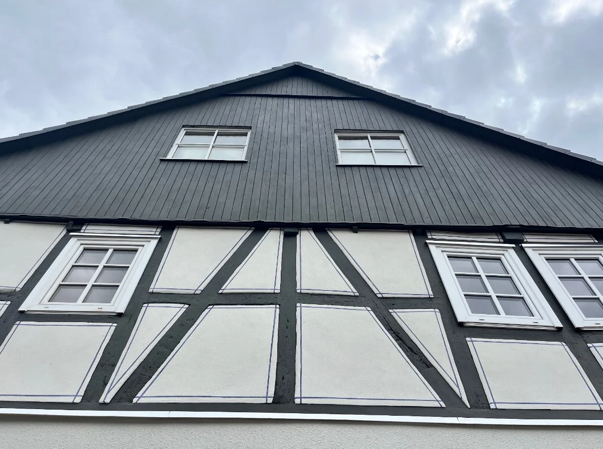Aufnahme eines Hauses im Fachwerkstil mit weißen Wänden und schwarzen Holzbalken, darunter befinden sich mehrere weiße Fenster, der Himmel ist teils bewölkt.