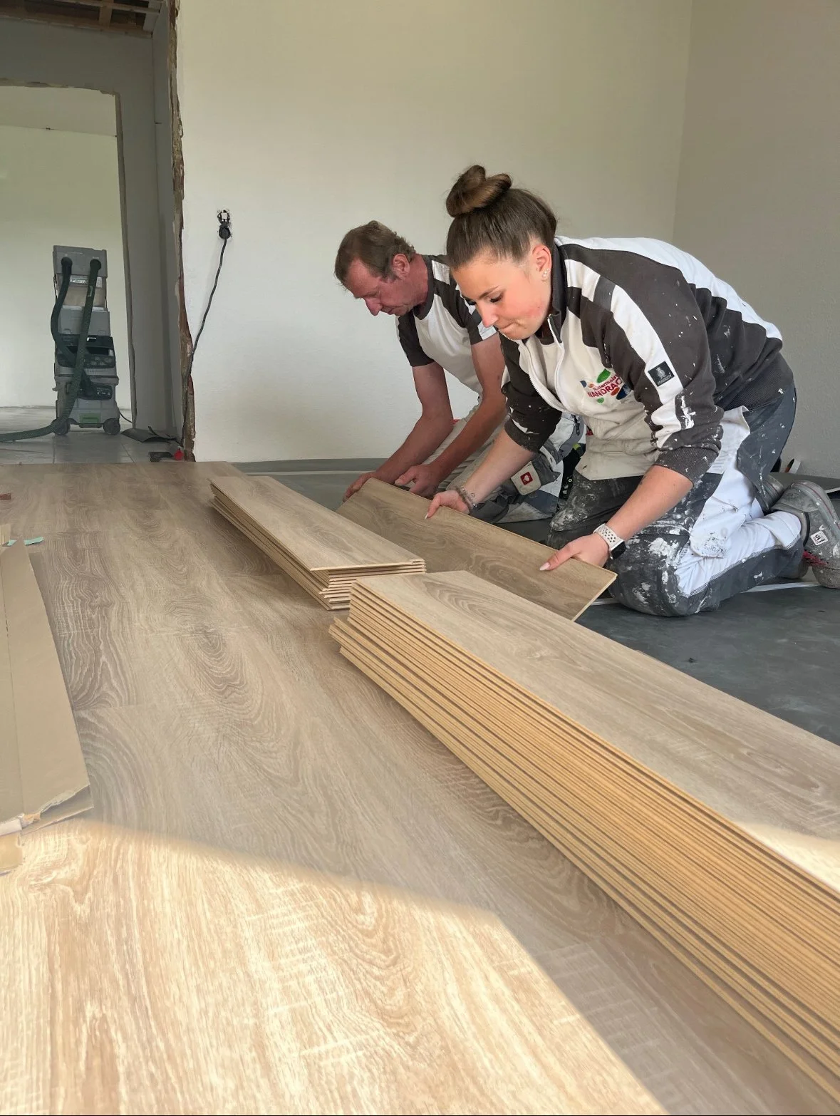 Zwei Frauen arbeiten an dem Verlegen von Holzdielen auf einem Boden, knien auf dem Boden, mit mehreren Holzstücken vor ihnen, in einem Raum, der renoviert wird.