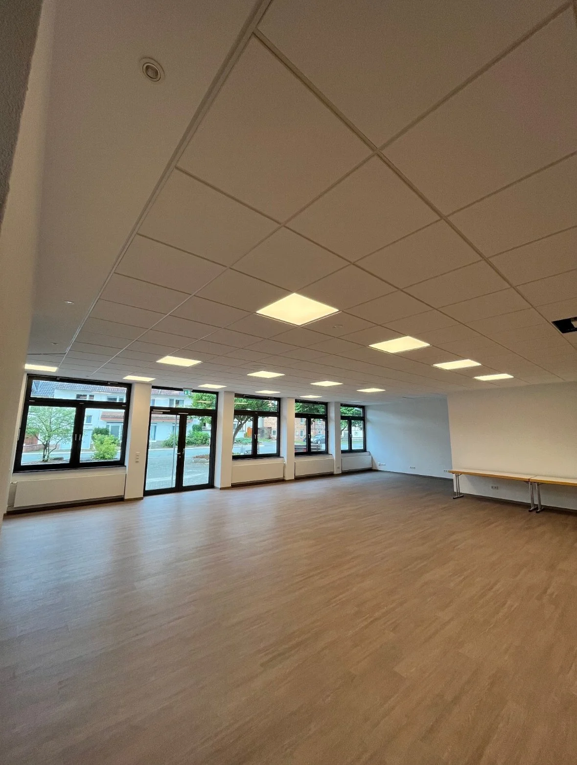 Leeres, gut beleuchtetes Zimmer mit großen Fenstern, Holzfußboden und weißen Wänden.