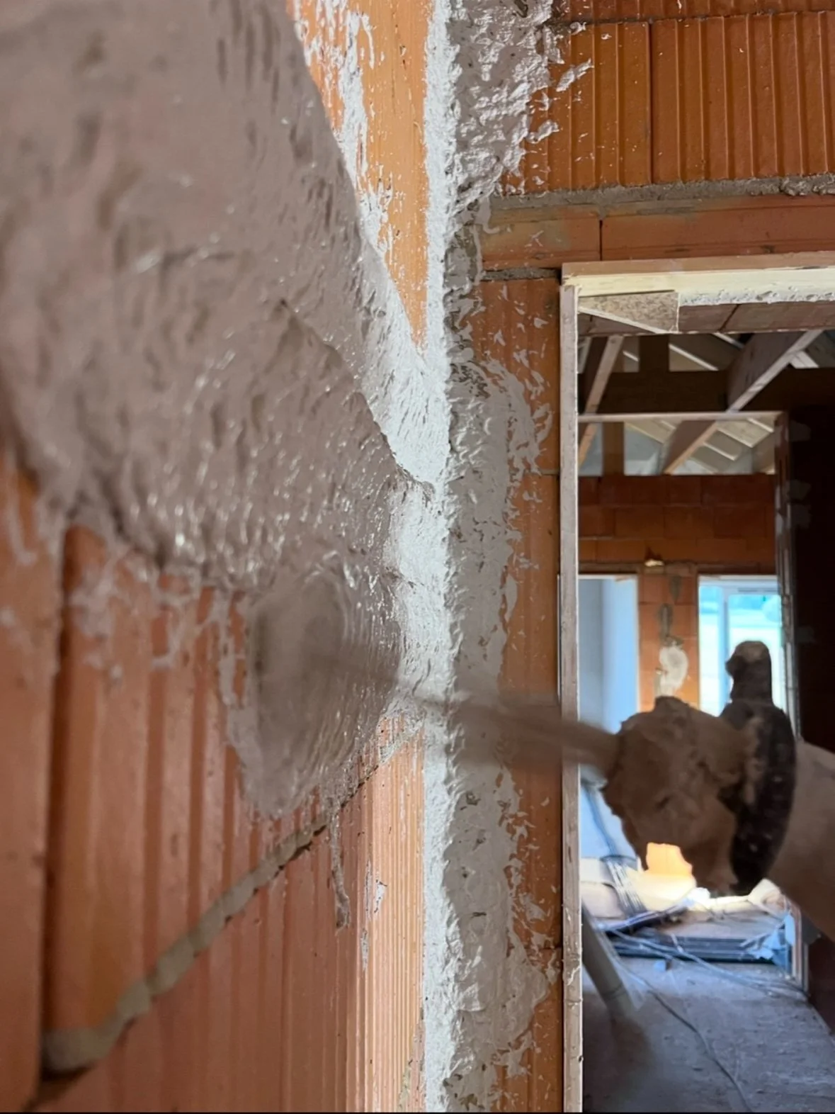Ein Handwerker trägt Mörtel an eine Wand in einem Innenraum auf, sichtbare Holzrahmen und Ziegelwände im Hintergrund.
