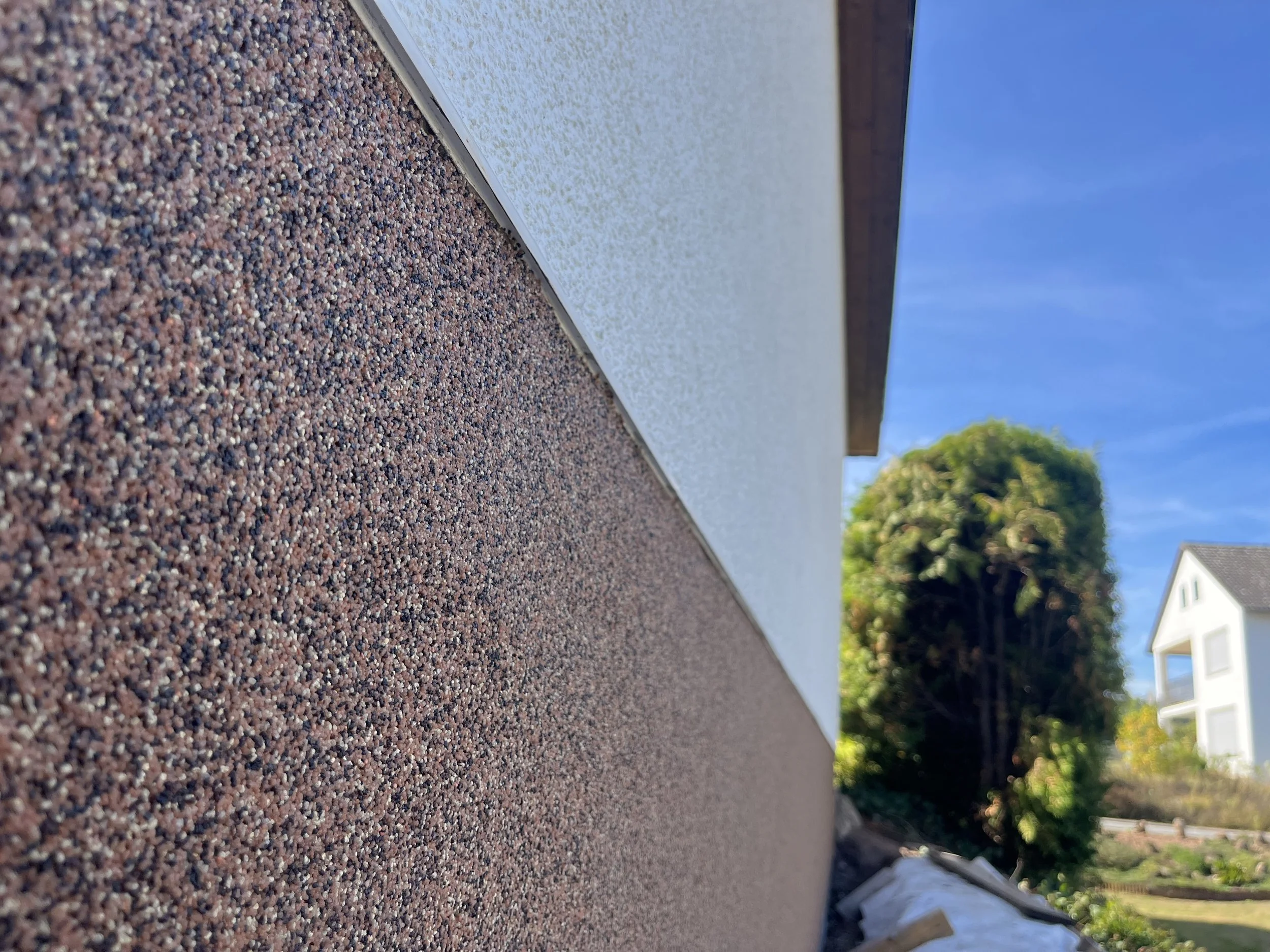 Nahaufnahme der Gebäudefassade mit grauer und roter Struktur, im Hintergrund ein blauer Himmel, Bäume und angrenzende Wohnhäuser.