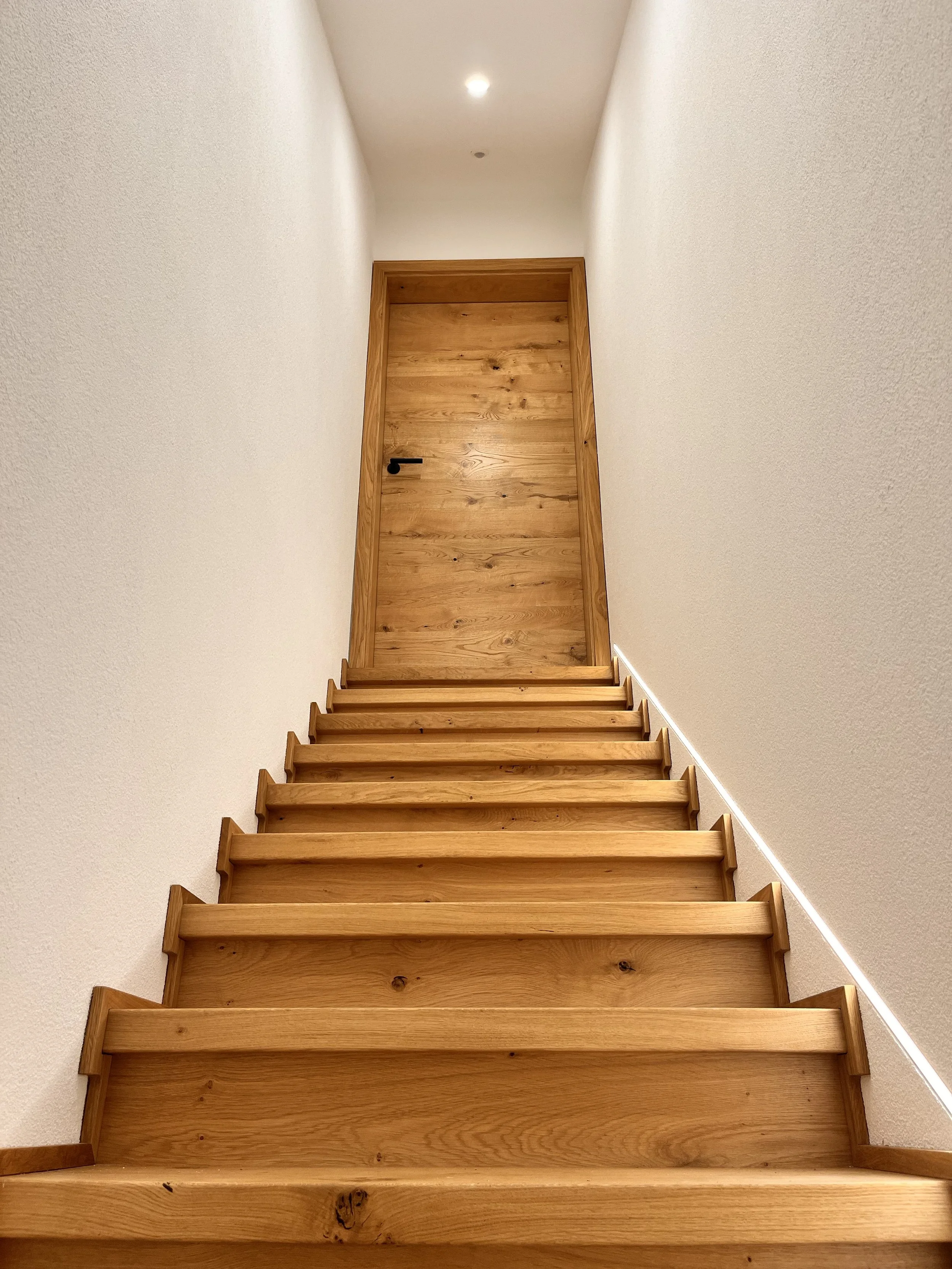 Holztreppe führt zu einer Tür in einem hellen, schmalen Flur.