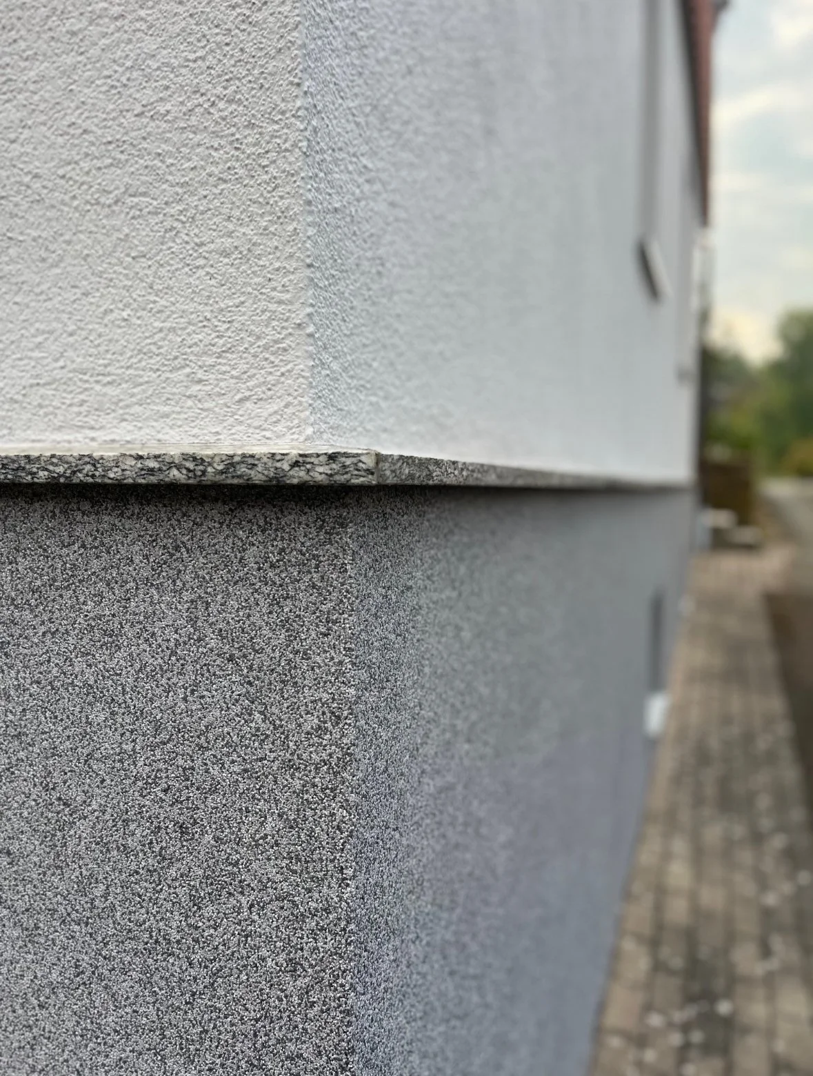 Nahaufnahme der Außenfassade eines Gebäudes mit einer hellgrauen Mauer und einer purpurroten Fensterbank, Blick seitlich entlang der Wand, unscharfer Hintergrund mit Gehweg.