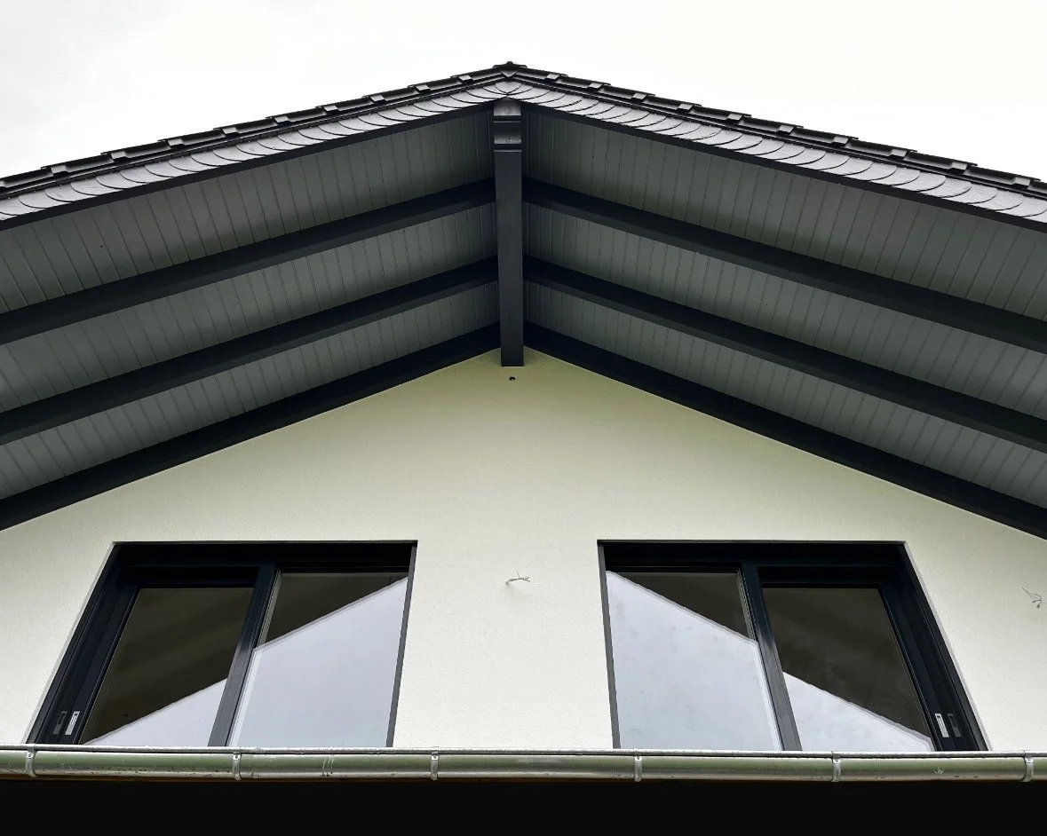 Ansicht eines modernen Hauses mit weißer Fassade, zwei große Fenster und einem schrägen Dach mit schwarzen Dachziegeln. 