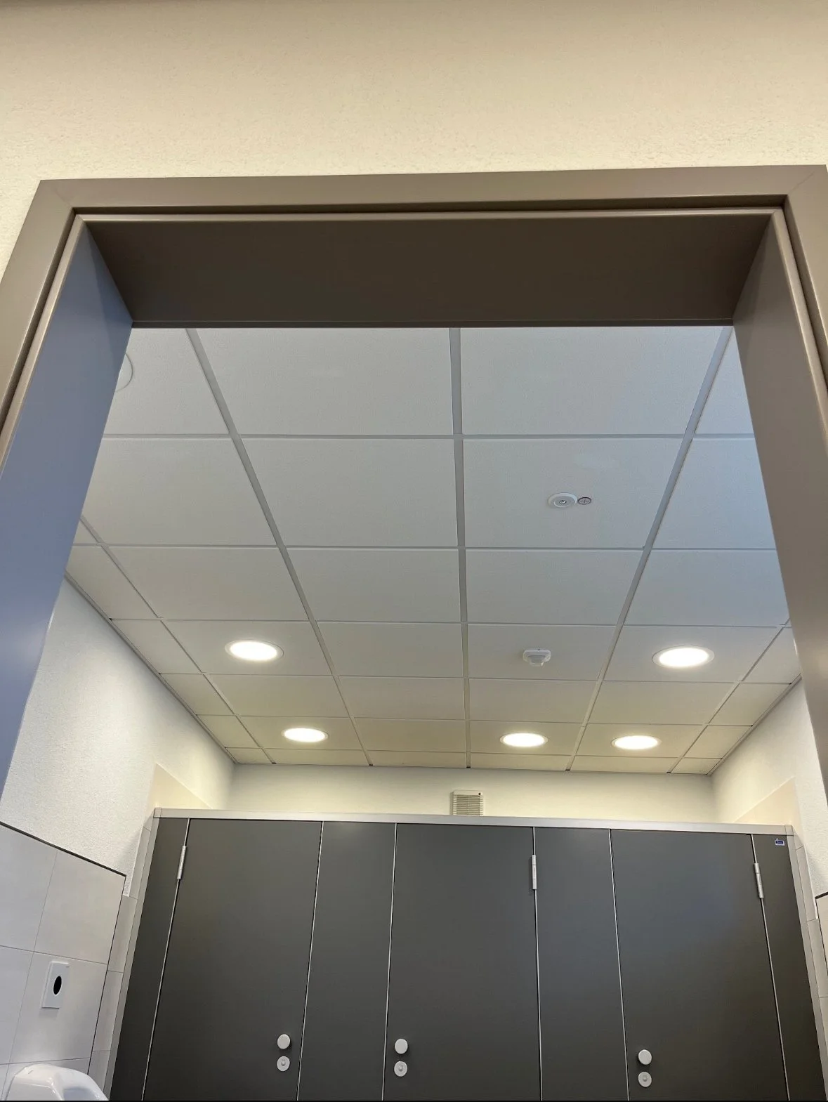 Innenaufnahme einer Umkleidekabine in einem öffentlichen Gebäude mit grauen Sperrholztüren und abgehängter Decke mit eingebauten Lichtleuchten.
